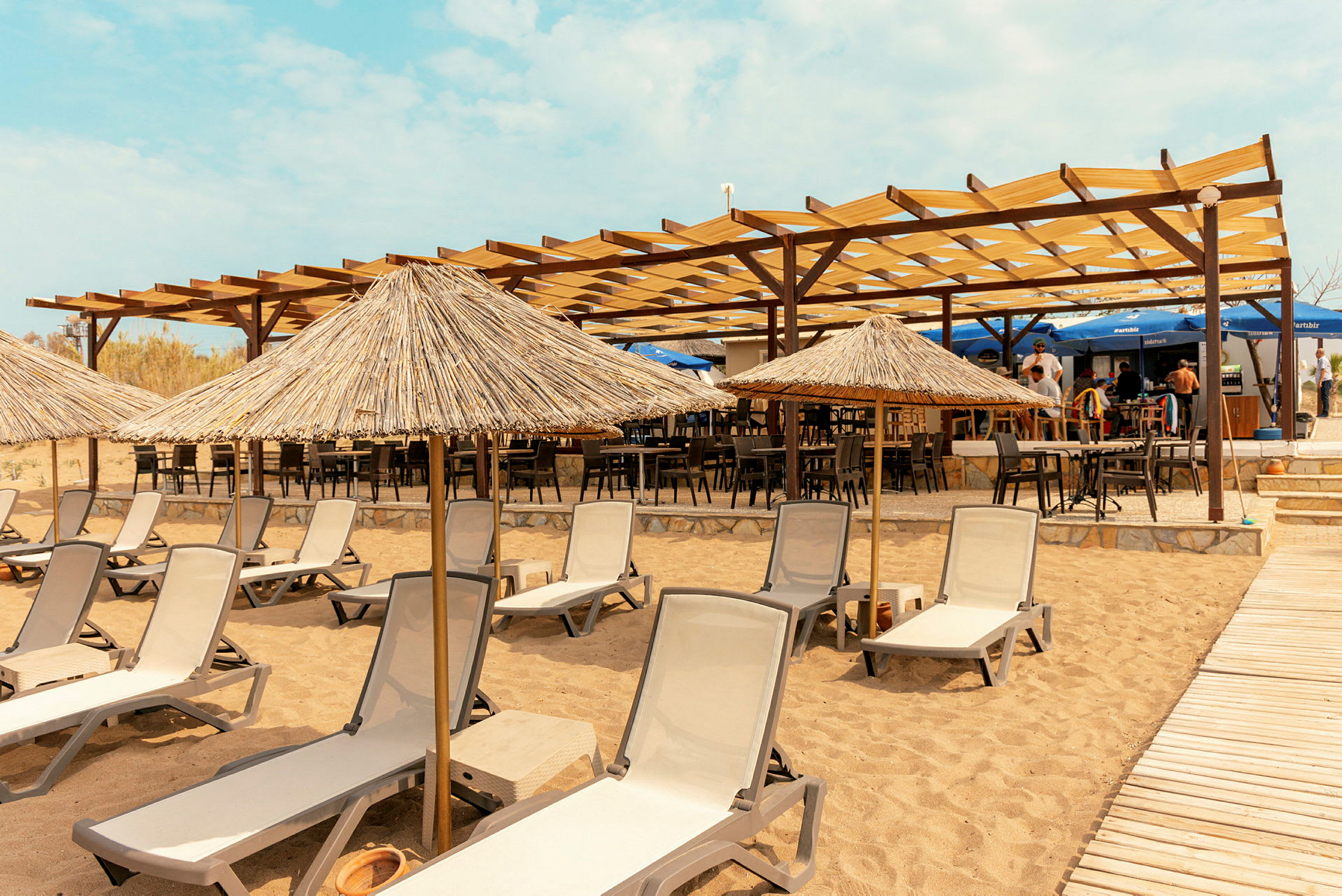 Strand med solstolar och halmtäckta parasoller framför en restaurang med öppet tak. (AI-genererad text)