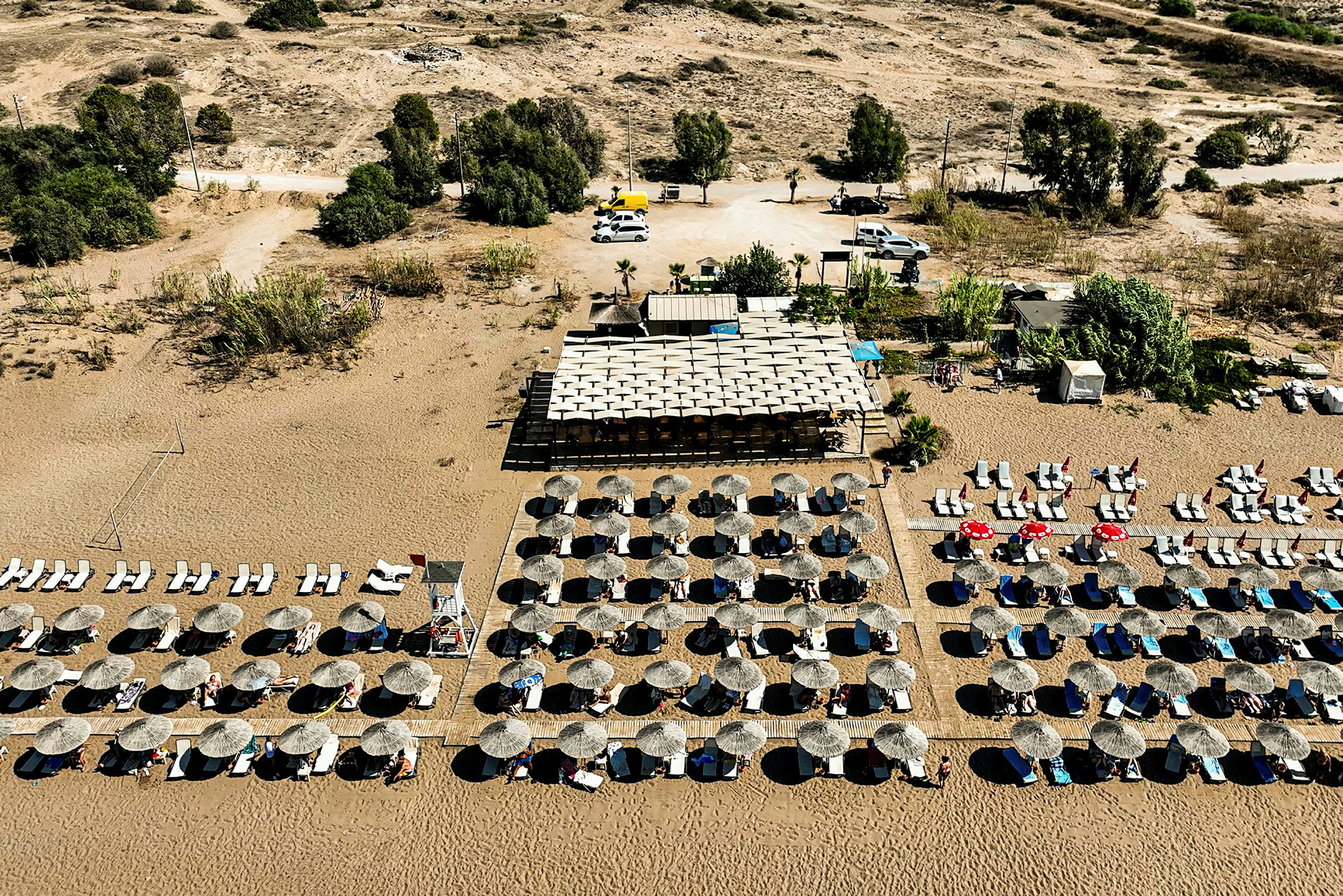 Flygfotografi av en strand med parasoller och solstolar på sanden samt en byggnad nära stranden. (AI-genererad text)