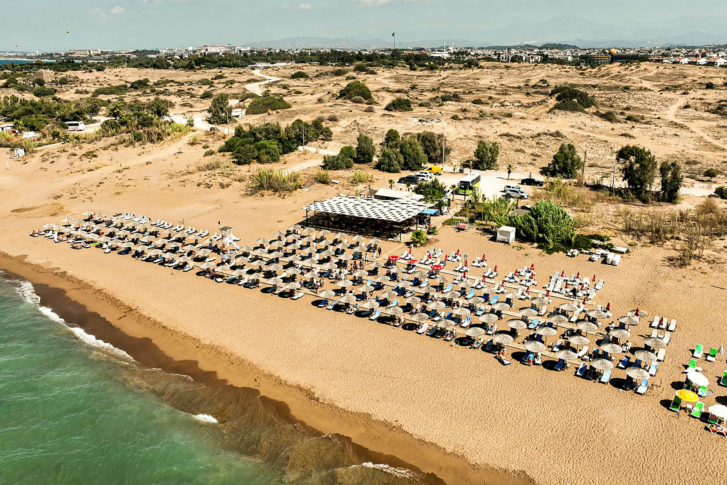 En lång sandstrand kantad med radvis av solstolar och parasoller med ett strandcafé i bakgrunden. (AI-genererad text)
