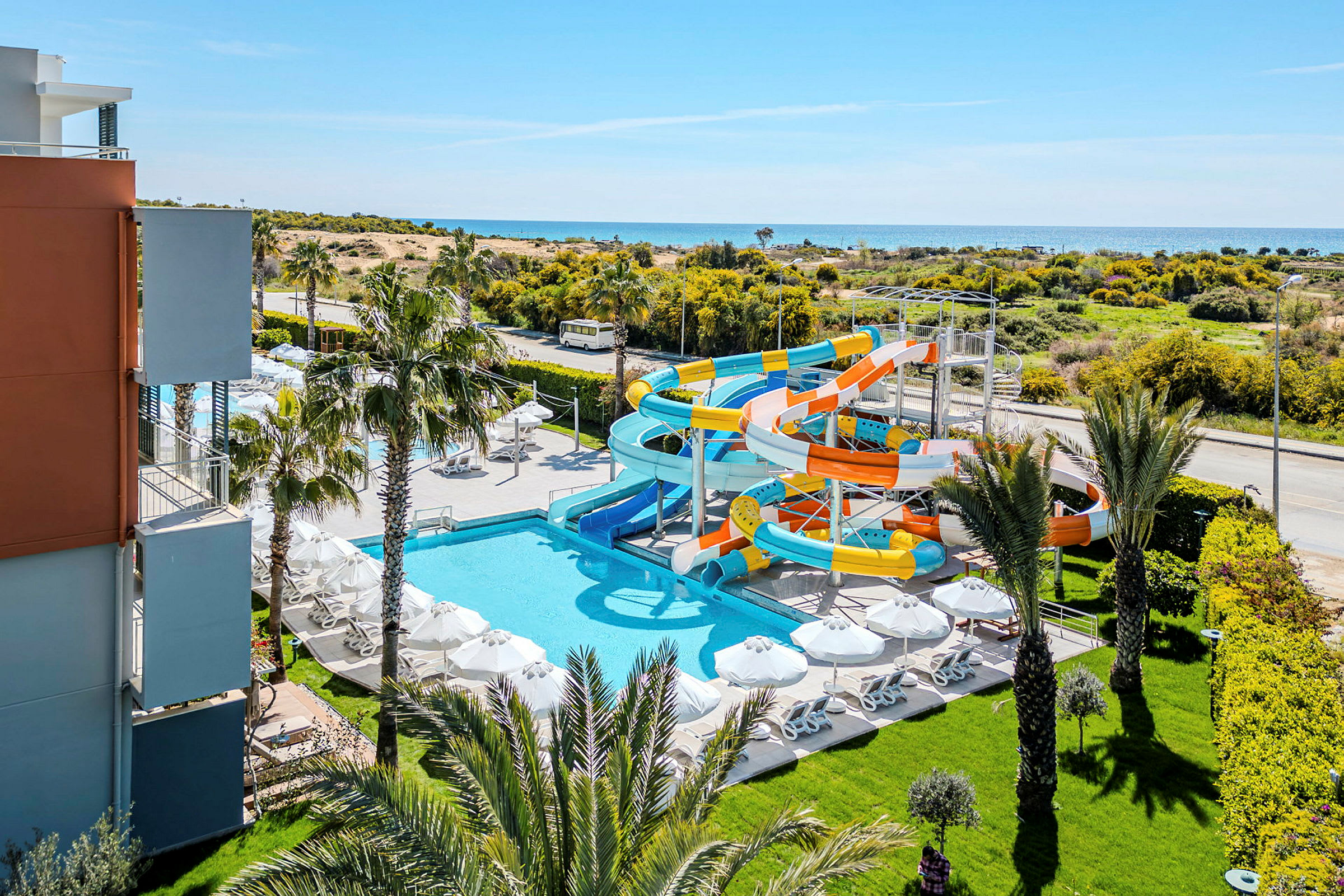 Bild av ett hotellområde med pool, solstolar och vattenrutschbanor, omgivning med träd och hav i bakgrunden. (AI-genererad text)