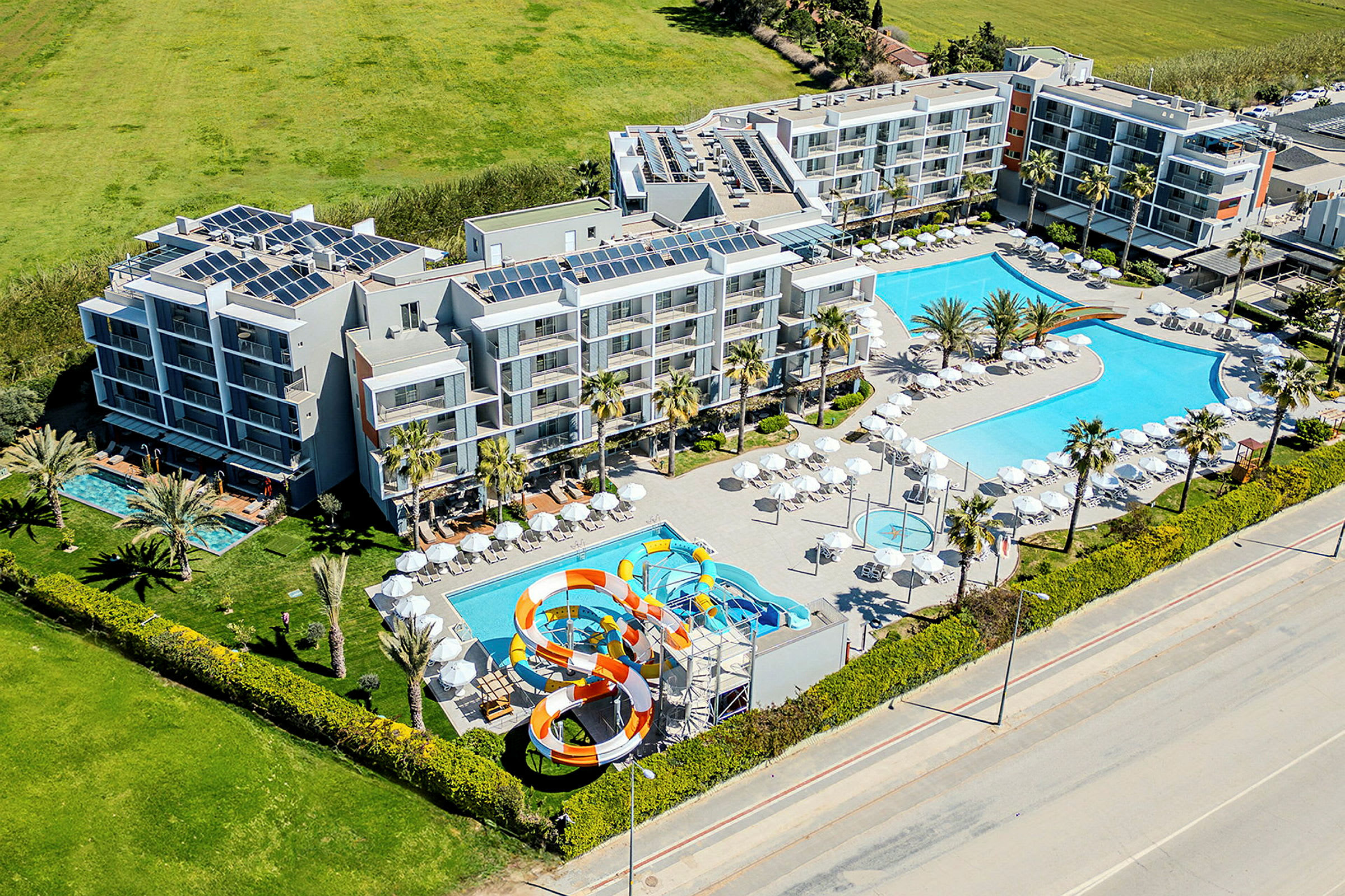 Hotellkomplex med flera byggnader, två stora poolområden och en vattenrutschkana, omgiven av grönska och en väg. (AI-genererad text)