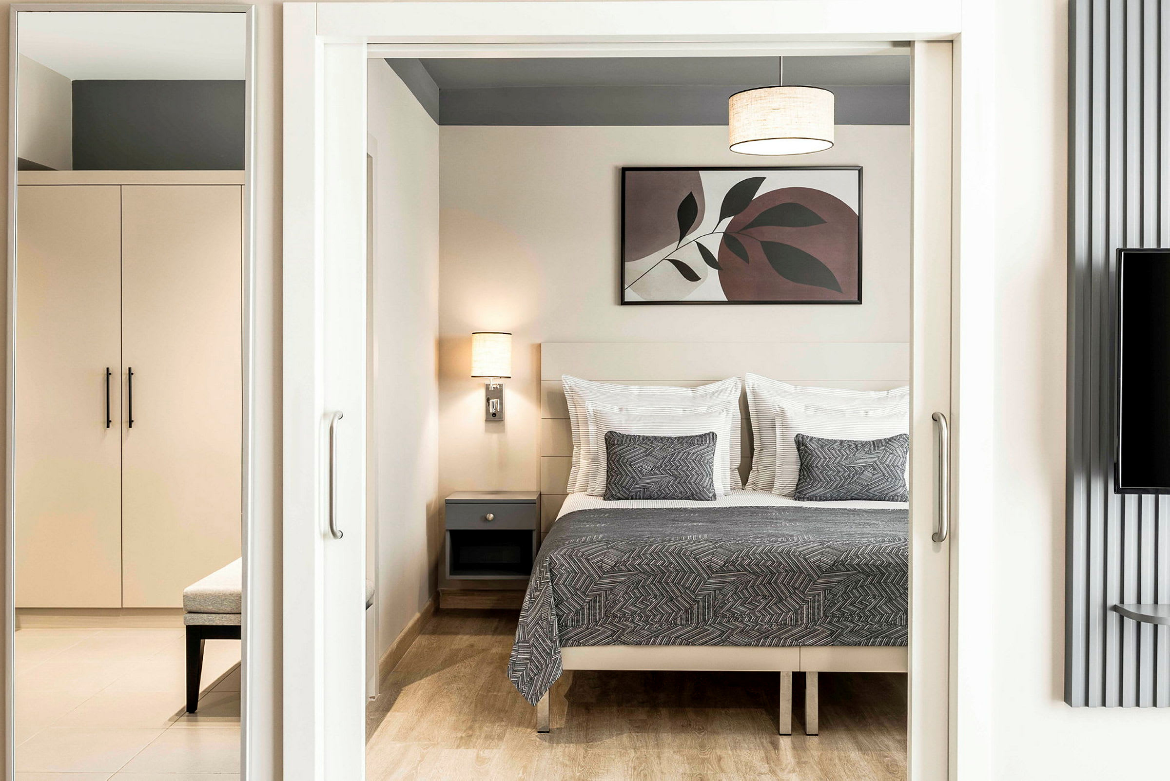 Ett sovrum med en dubbelbädd med grå quilt och matchande kuddar, en vägglampa, och en tavla ovanför sängen. Ett skåp och bänk syns i förgrunden. (AI-genererad text)