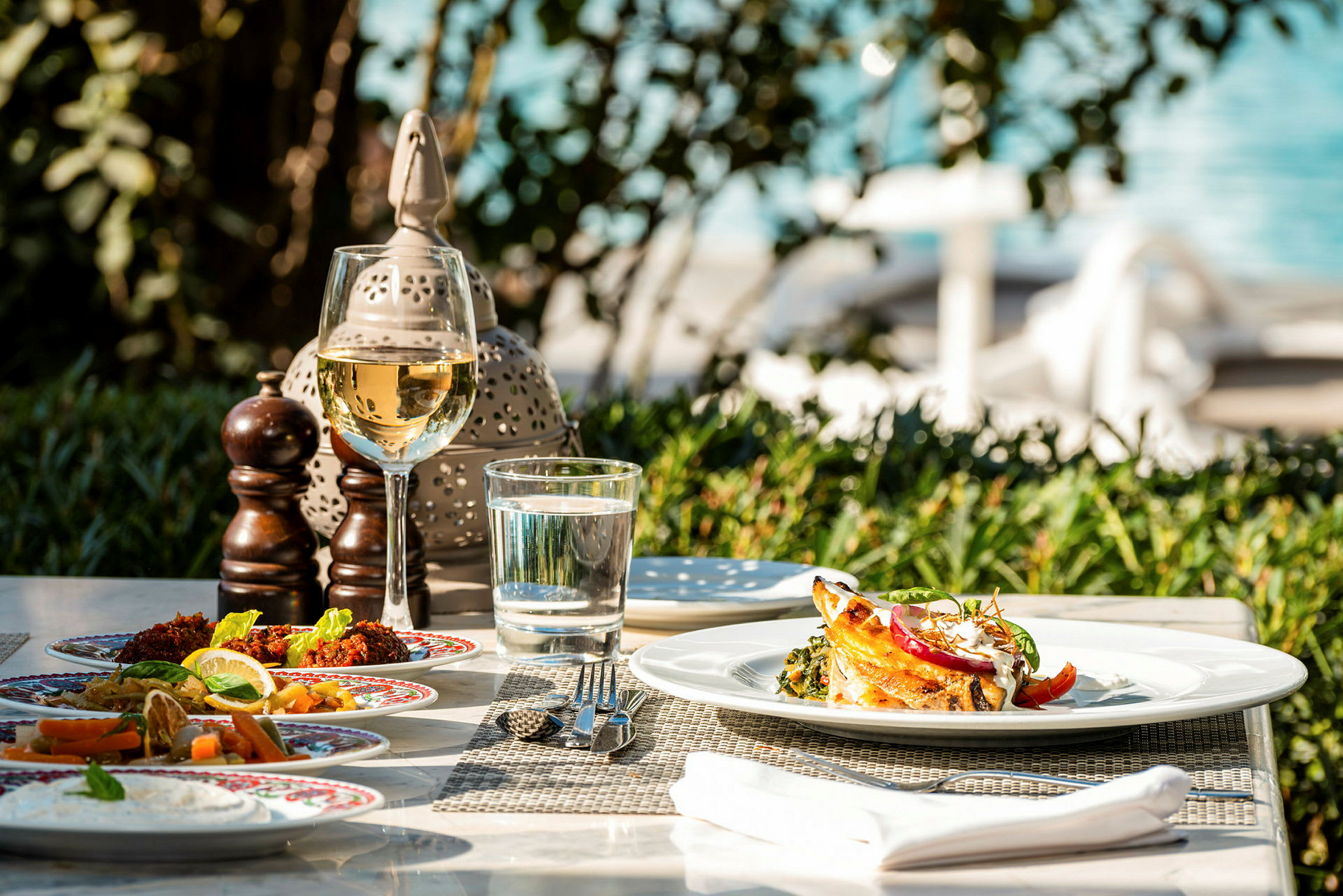 Ett dukat bord med tallrikar av olika maträtter, ett glas vin och en vattenglas vid utomhusmiljö. (AI-genererad text)