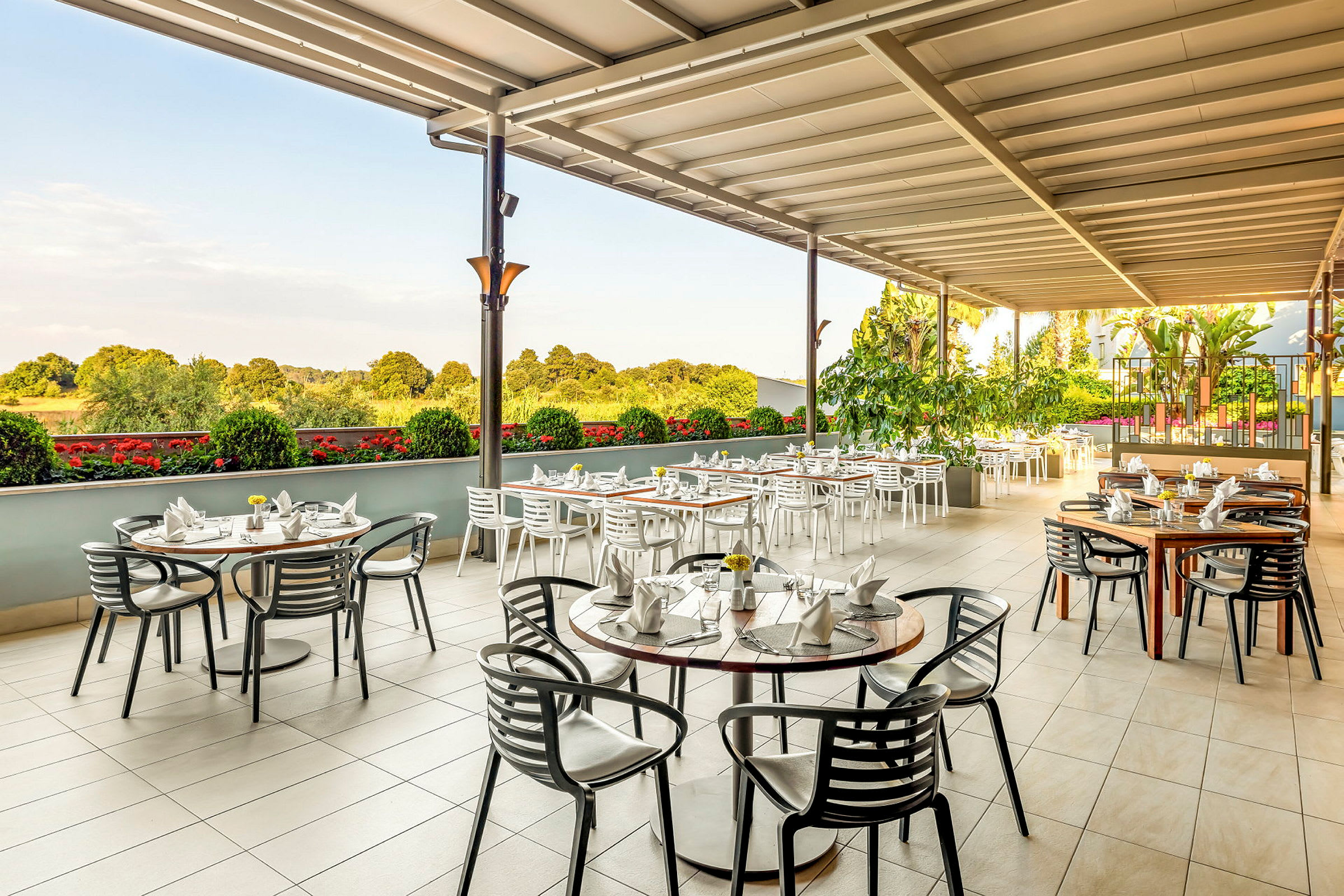 Utomhusrestaurang med dukade bord och stolar under tak, omgiven av gröna växter. (AI-genererad text)