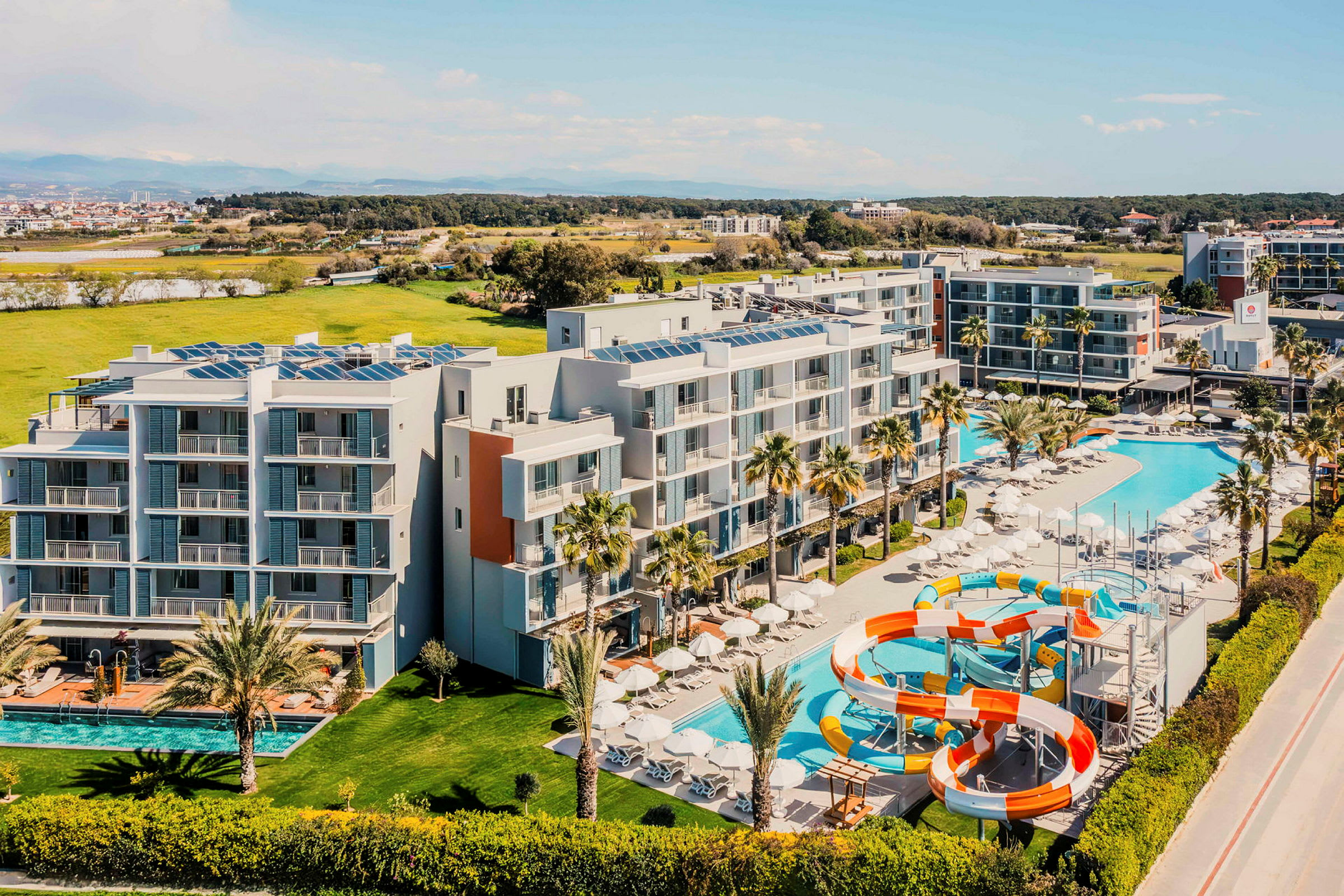 Flygfoto av ett hotell med pooler och vattenrutschbanor, omgiven av grönområden. (AI-genererad text)