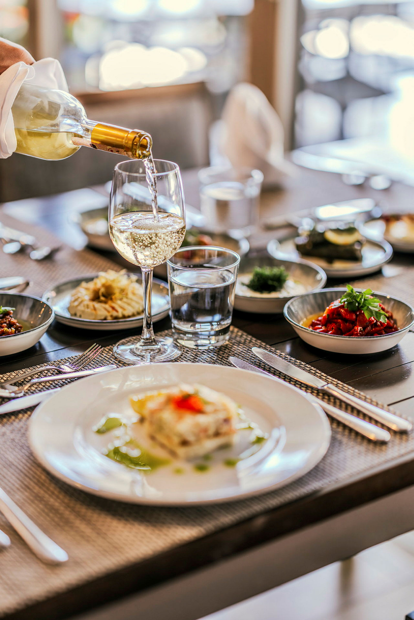 En hand häller vitt vin i ett vinglas på ett dukat bord med mat. (AI-genererad text)