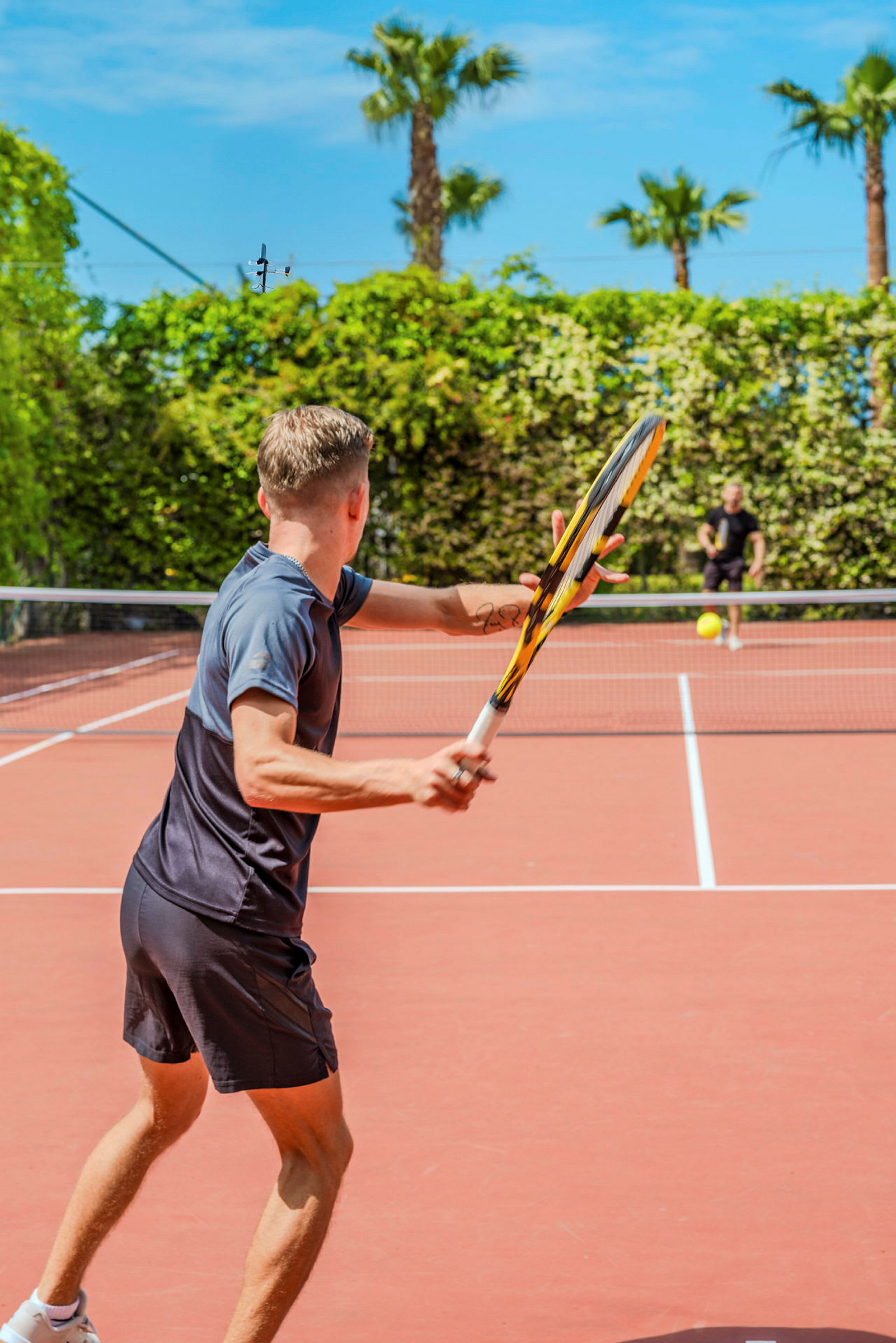 Två personer spelar tennis utomhus på en tennisbana omgiven av grönska och palmer. (AI-genererad text)