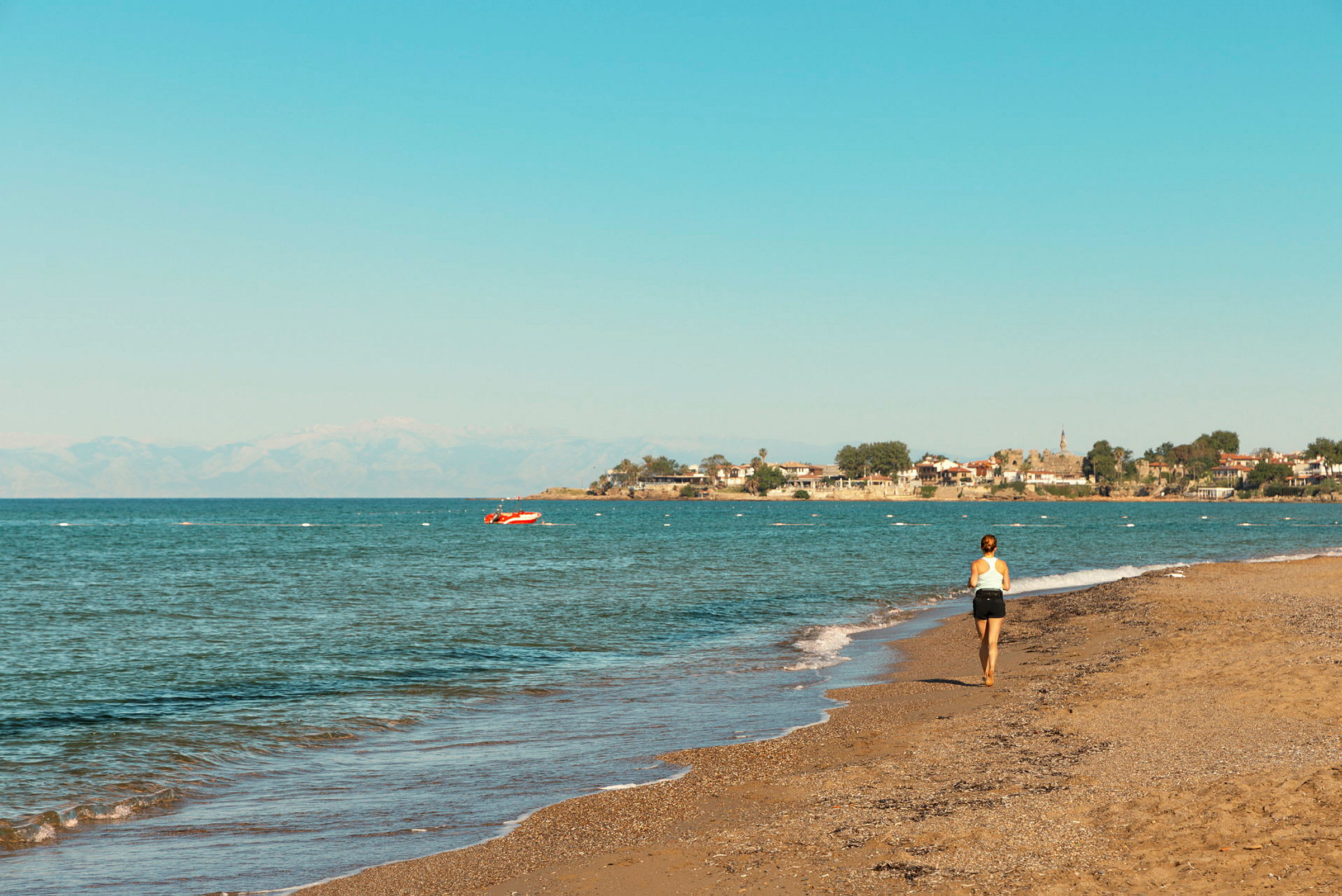 En kvinna går längs en strand med utsikt över havet och en by vid kusten i bakgrunden. (AI-genererad text)