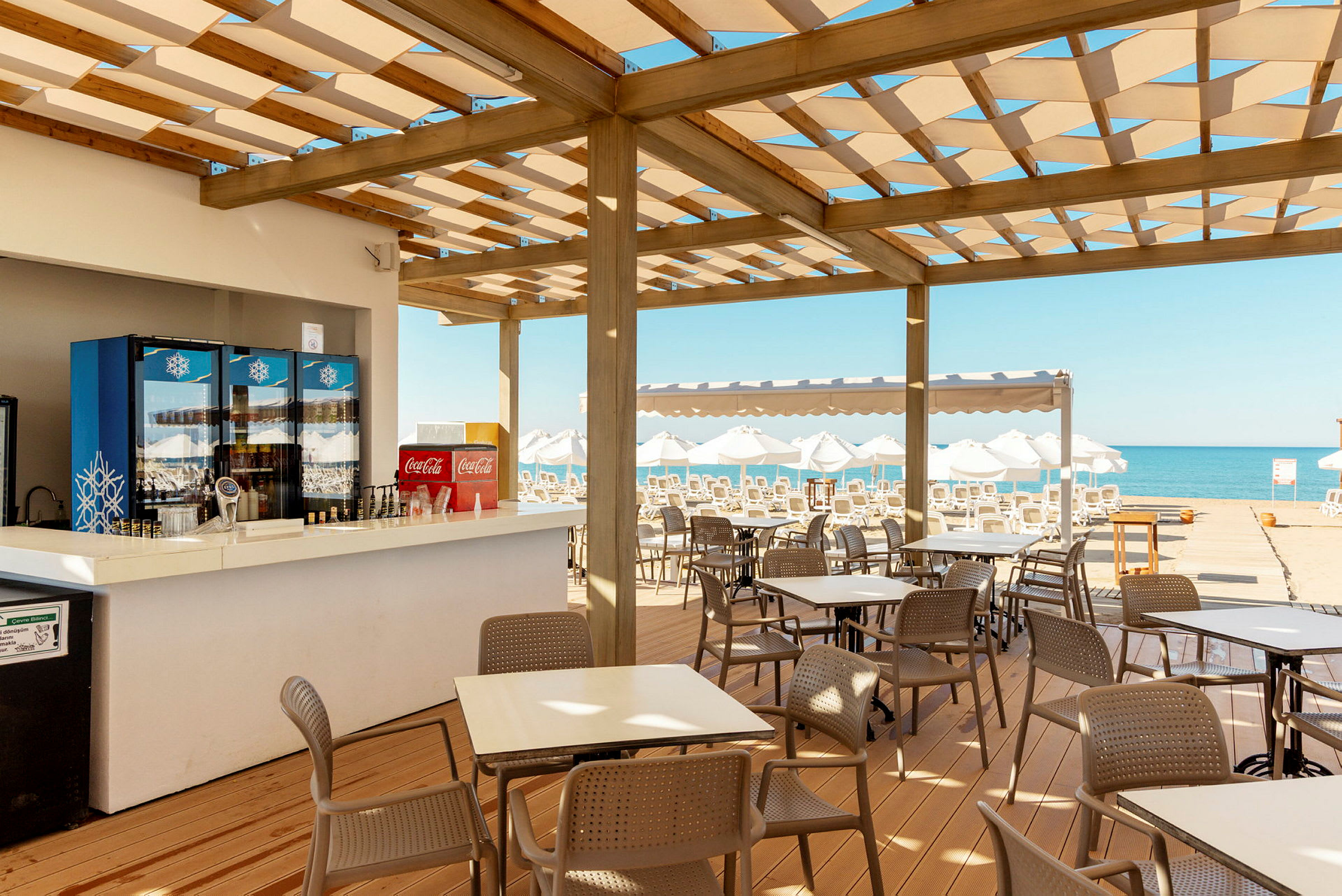 Ett kafé med uteservering under ett trätak nära stranden. Bord och stolar står utplacerade, och i bakgrunden syns solstolar och havet. (AI-genererad text)