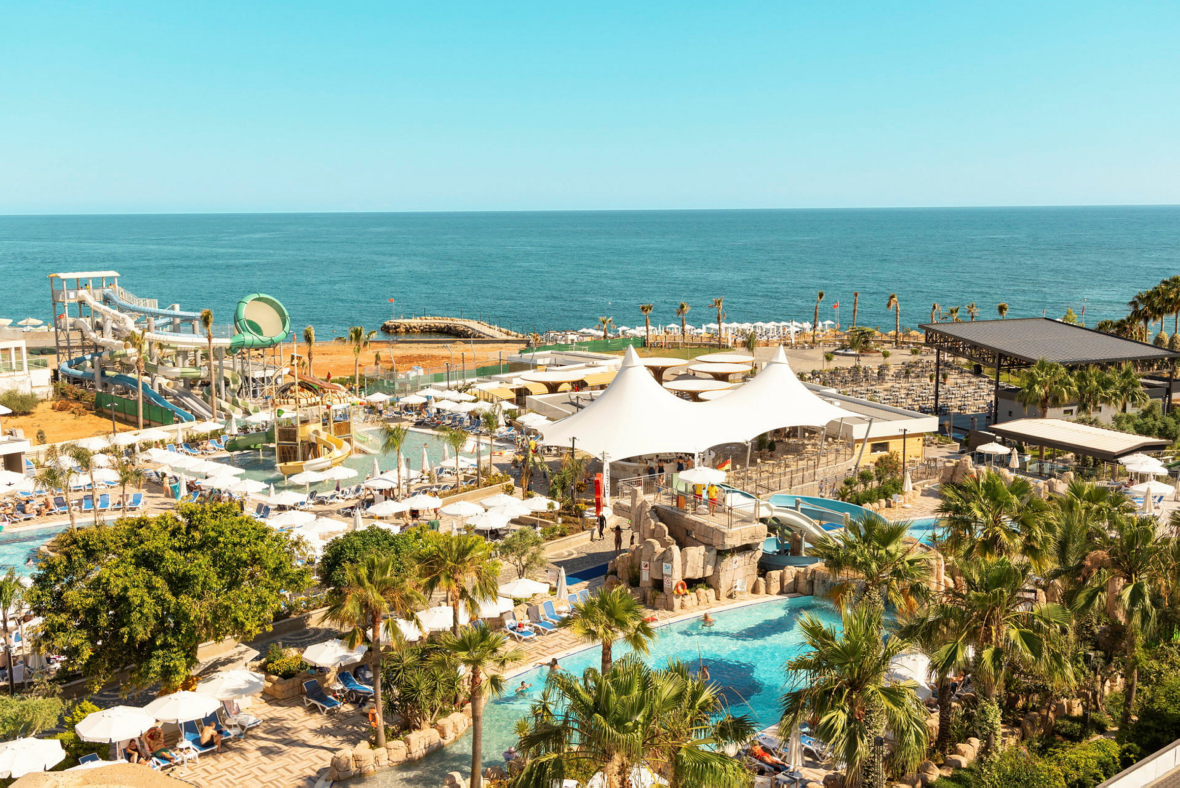 Vy över ett semesterresort vid havet med pool, vattenrutschbanor och palmer. (AI-genererad text)