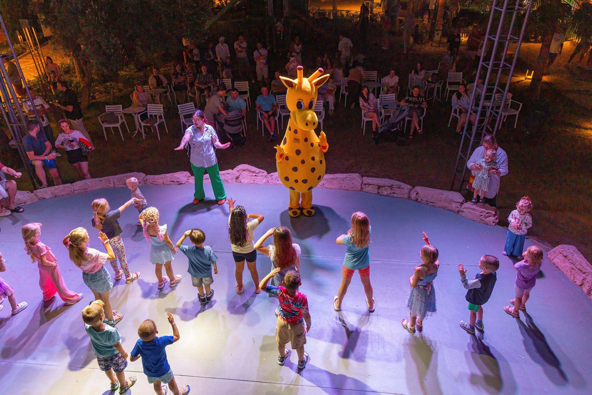 En grupp barn dansar runt en maskot föreställande en gul giraff på en scen, medan vuxna sitter och tittar i bakgrunden. (AI-genererad text)