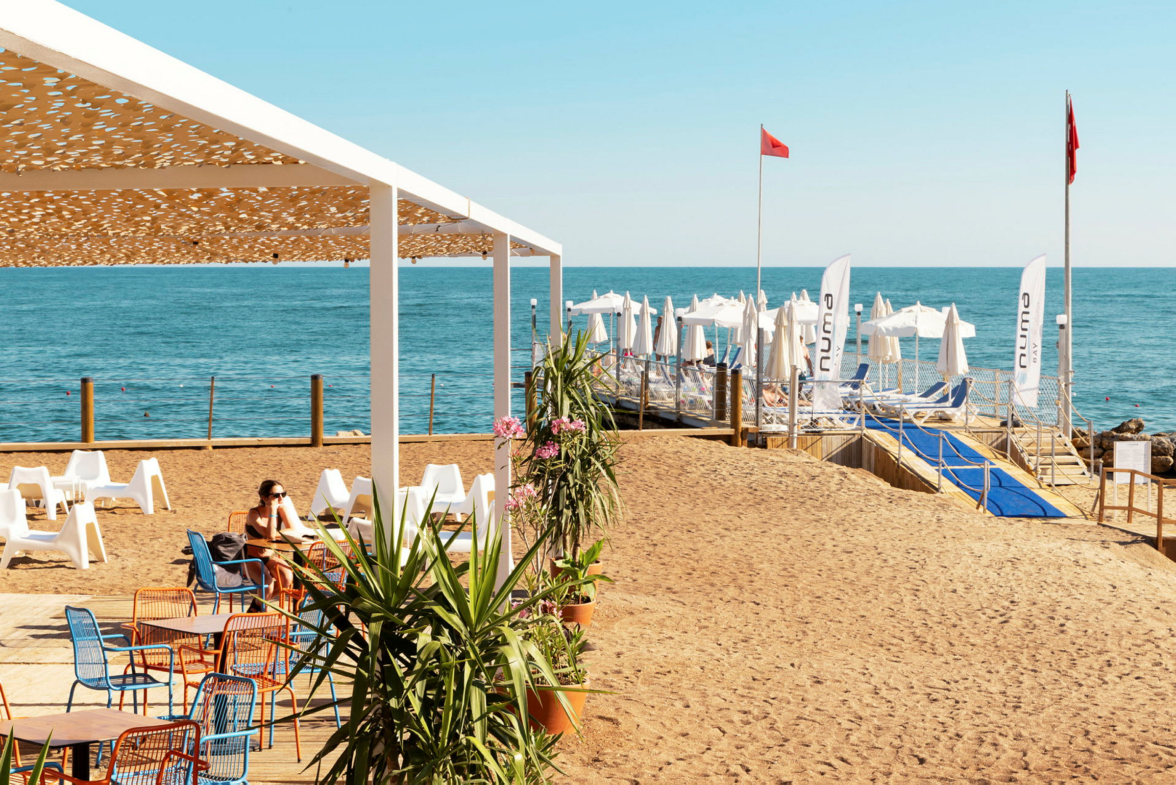 En strand med stolar och bord under en pergola, och i bakgrunden ses solstolar och parasoll vid havet. (AI-genererad text)