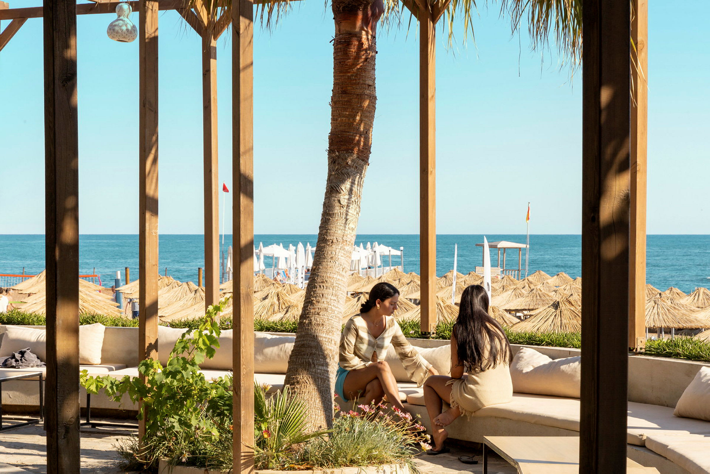 Två personer sitter på en utomhussoffa under ett träpergola och tittar ut mot havet och stranden med parasoller. (AI-genererad text)
