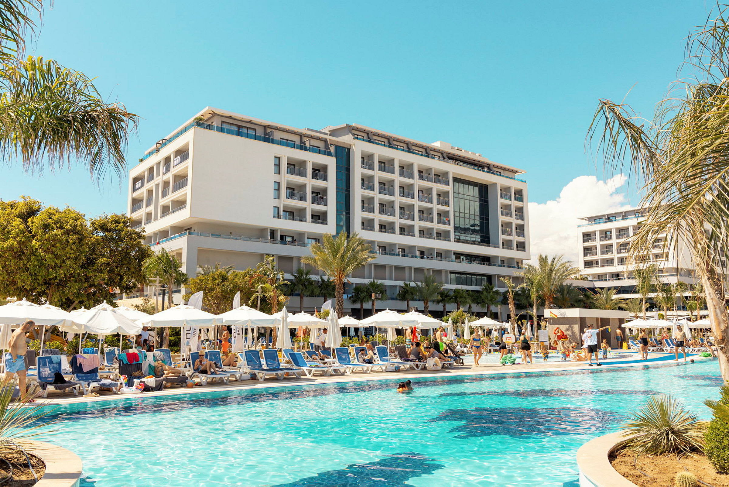 En stor semesteranläggning med pool, solstolar och parasoller i förgrunden och en modern byggnad i bakgrunden. (AI-genererad text)