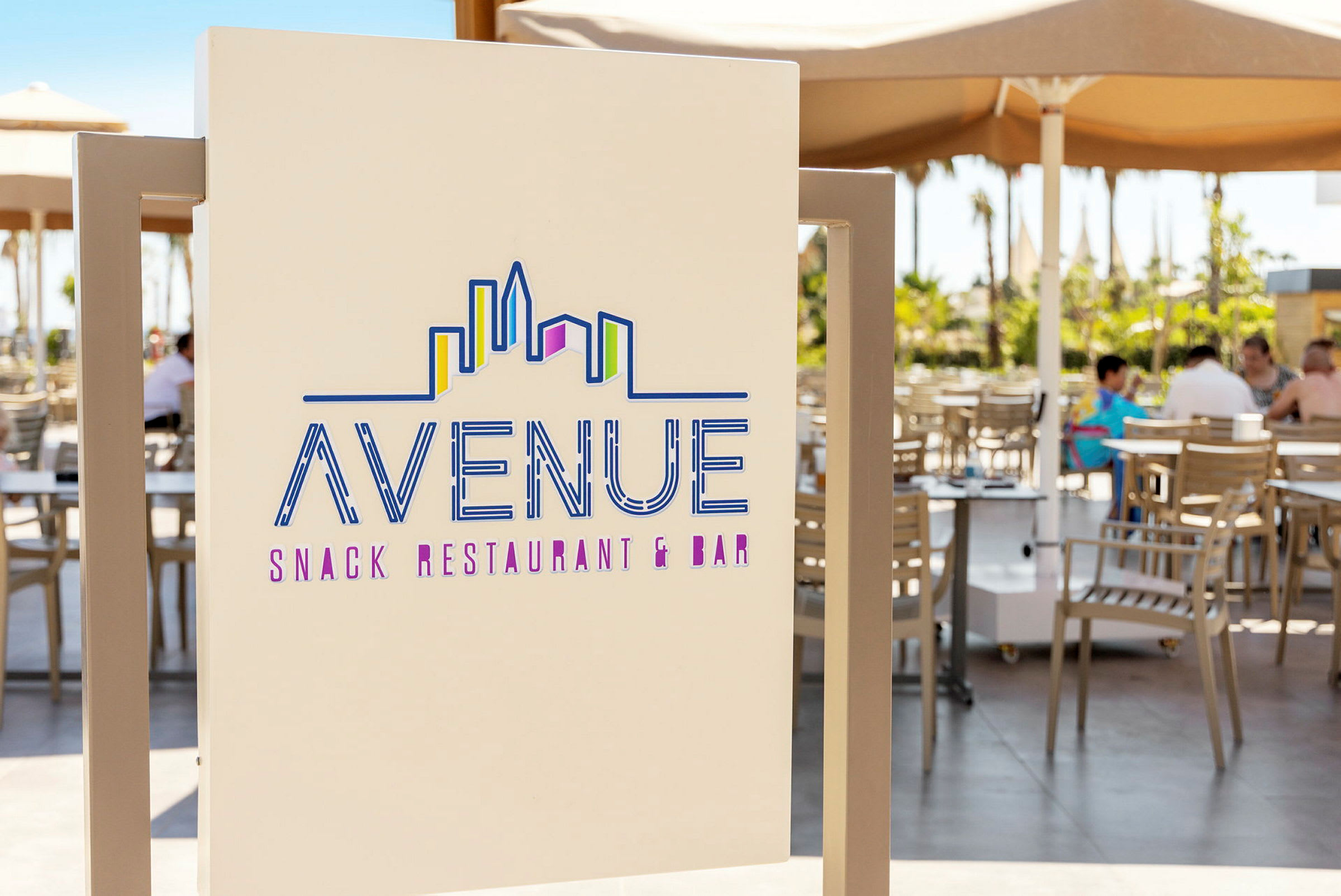 En skylt som visar texten "Avenue Snack Restaurant & Bar" med ett stiliserat skyline-motiv. I bakgrunden syns en uteservering med bord och stolar under parasoller. (AI-genererad text)