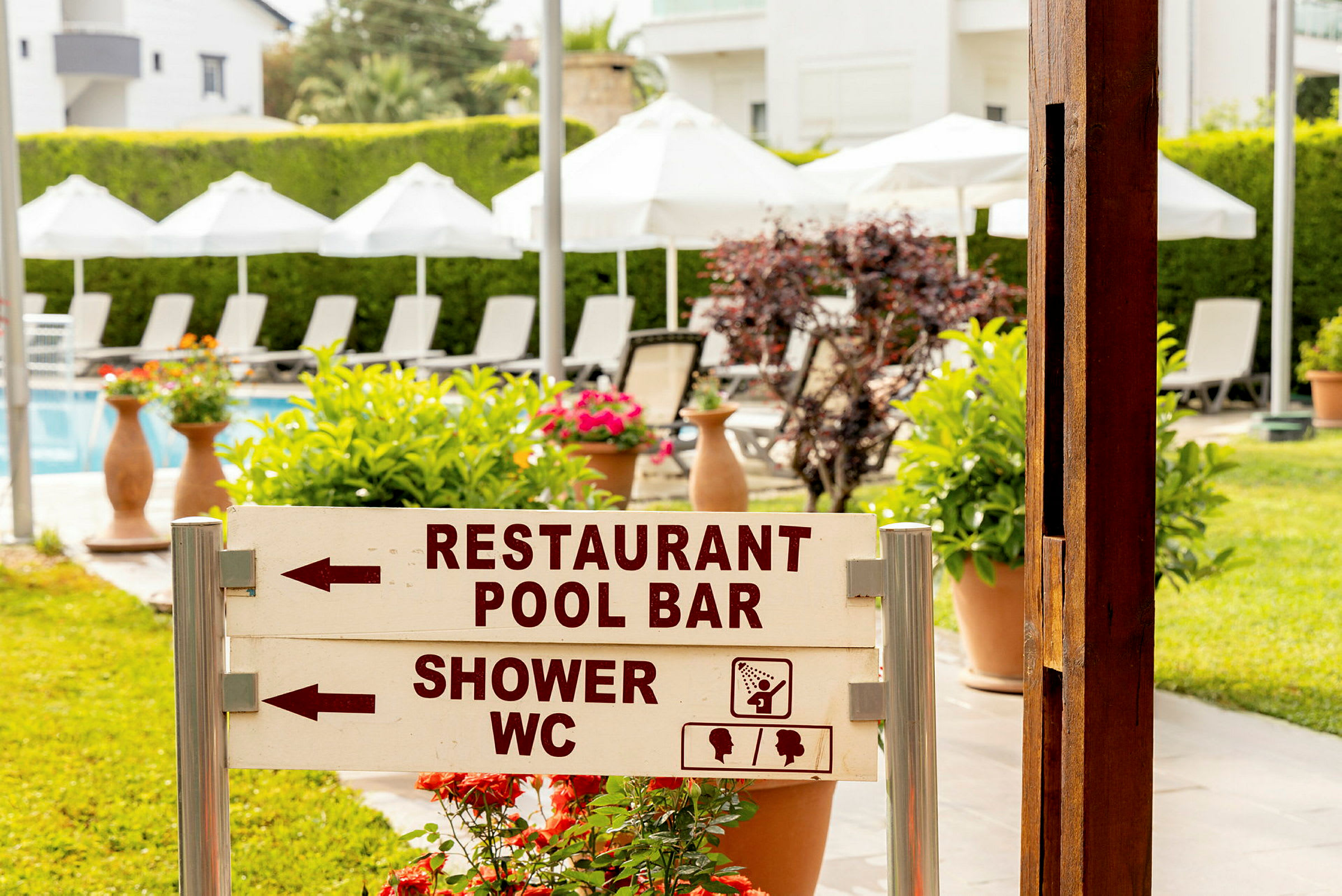 Skylt vid en trädgård med texten 'Restaurant Pool Bar' och 'Shower WC', solstolar och parasoller syns i bakgrunden. (AI-genererad text)