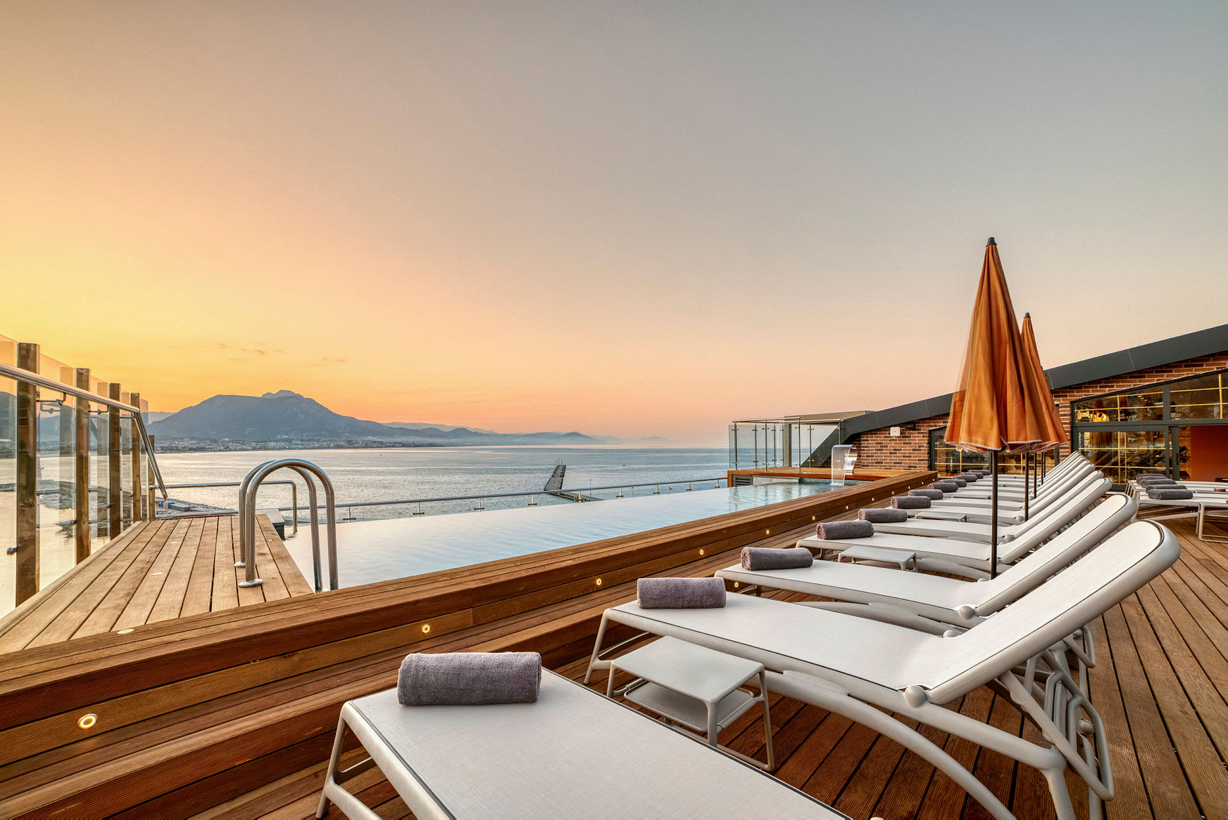 Solstolar och paraplyer vid en infinitypool med hav och berg i bakgrunden vid solnedgång. (AI-genererad text)