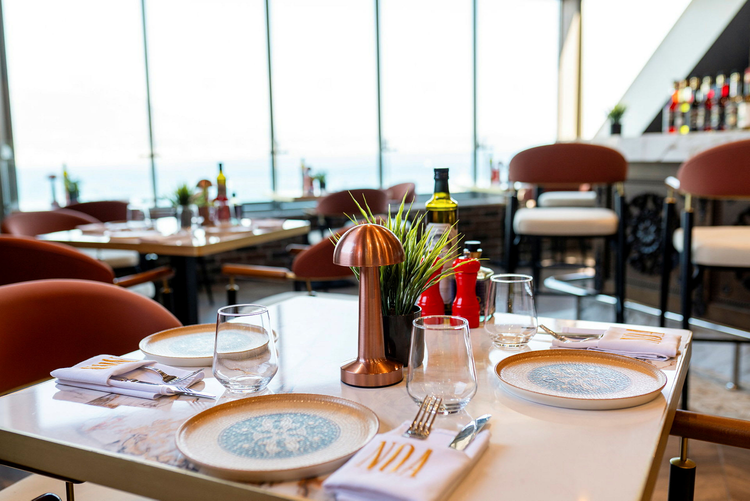 Bord på en restaurang med tallrikar, glas och bestick, utsikt mot stora fönster. (AI-genererad text)