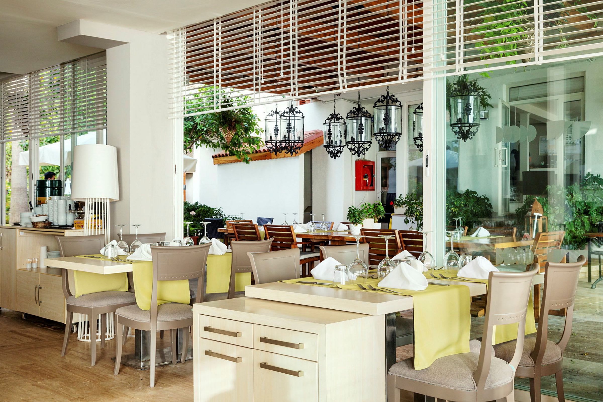 Restaurang med dukade bord och glasutsikt mot en terass med stolar och växter. (AI-genererad text)