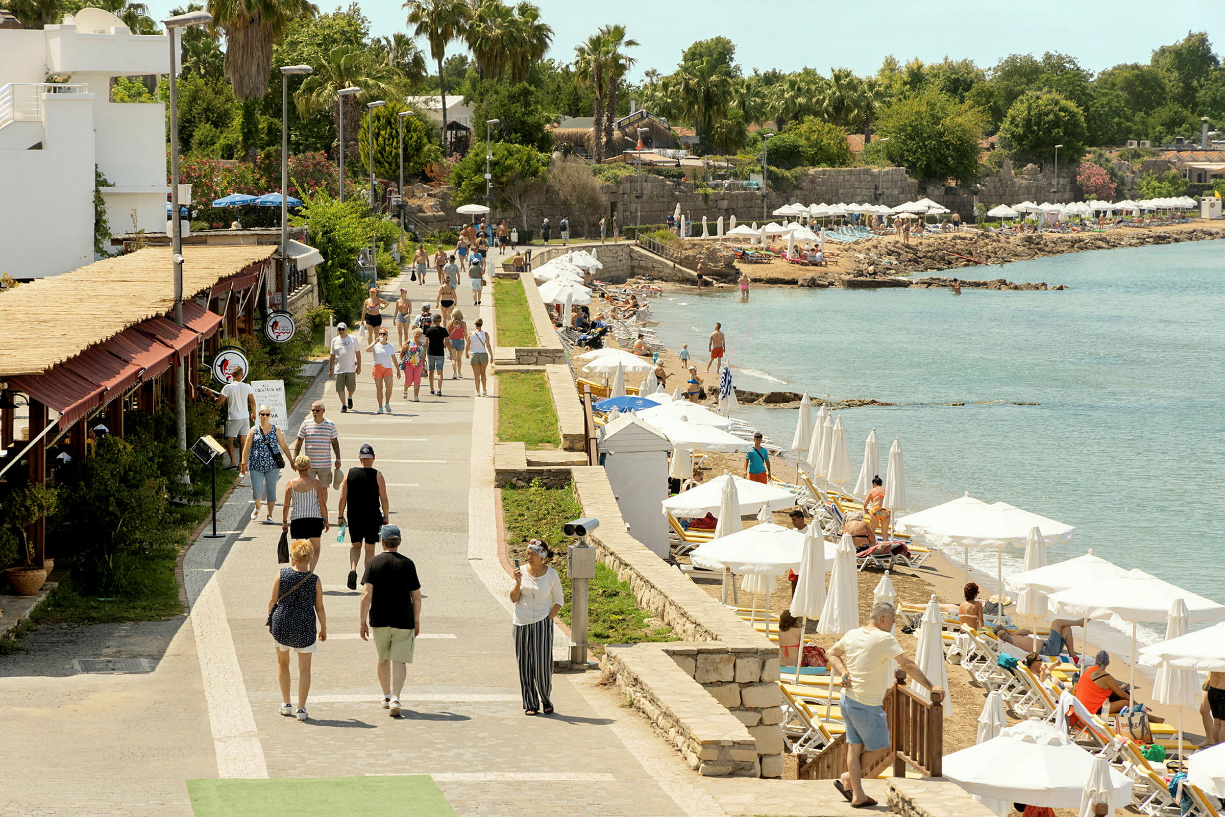En gångväg vid havet med människor som promenerar och personer som kopplar av på solstolar under parasoller. (AI-genererad text)