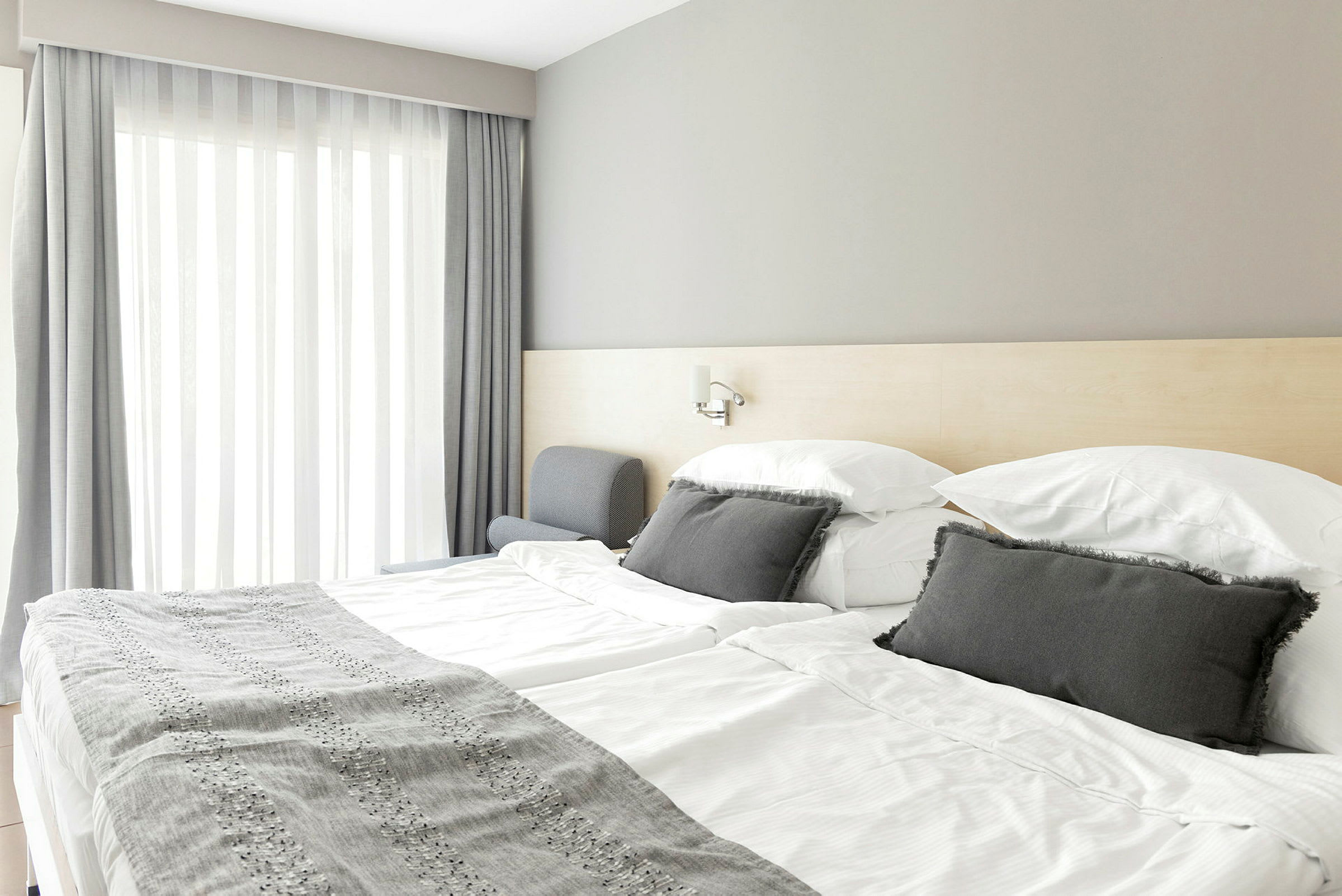 Ett hotellrum med en dubbelsäng, ljusa lakan och grå prydnadskuddar. (AI-genererad text)