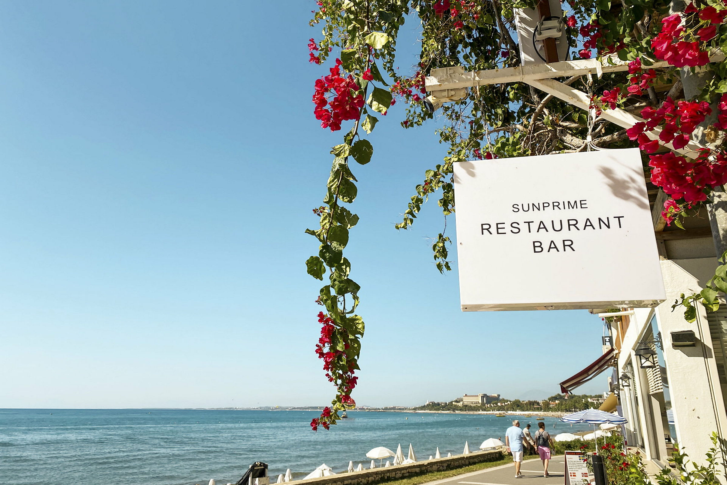 Skylt för Sunprime Restaurant Bar vid en promenad nära havet. (AI-genererad text)