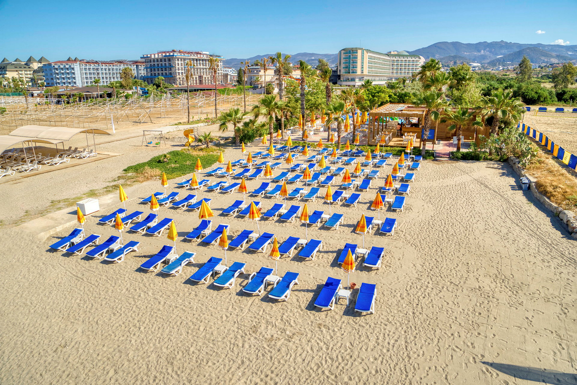 Solstolar och parasoller uppställda på en sandstrand med hotell och grönområden i bakgrunden. (AI-genererad text)