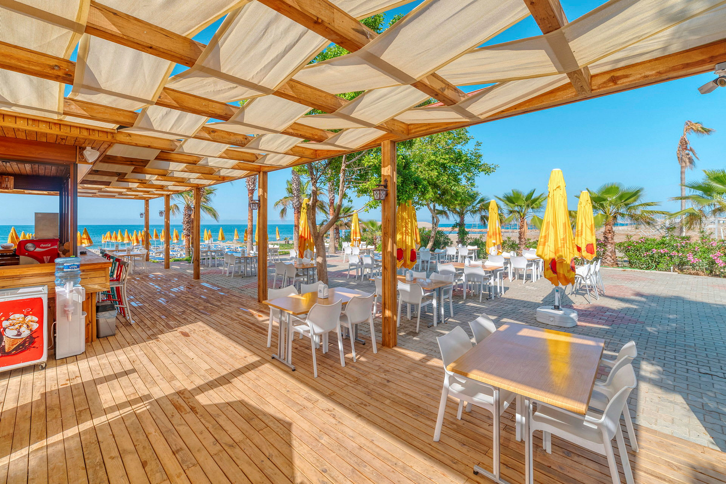 Utomhus matsal med bord och stolar under en pergola bredvid stranden. (AI-genererad text)