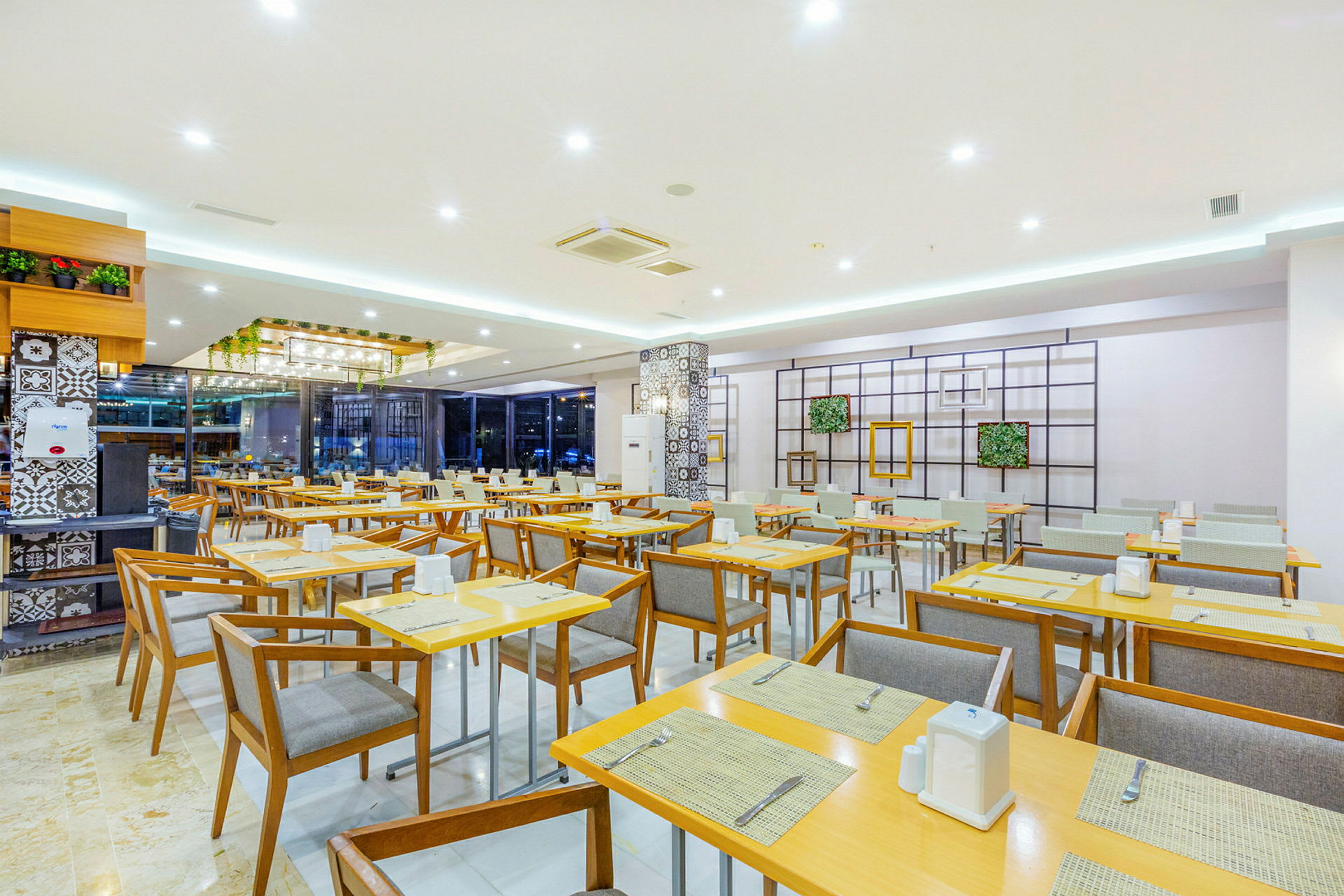 Ett rymligt restaurangområde med moderna trästolar och bord, ljus inredning och dekorativa växtarrangemang. (AI-genererad text)