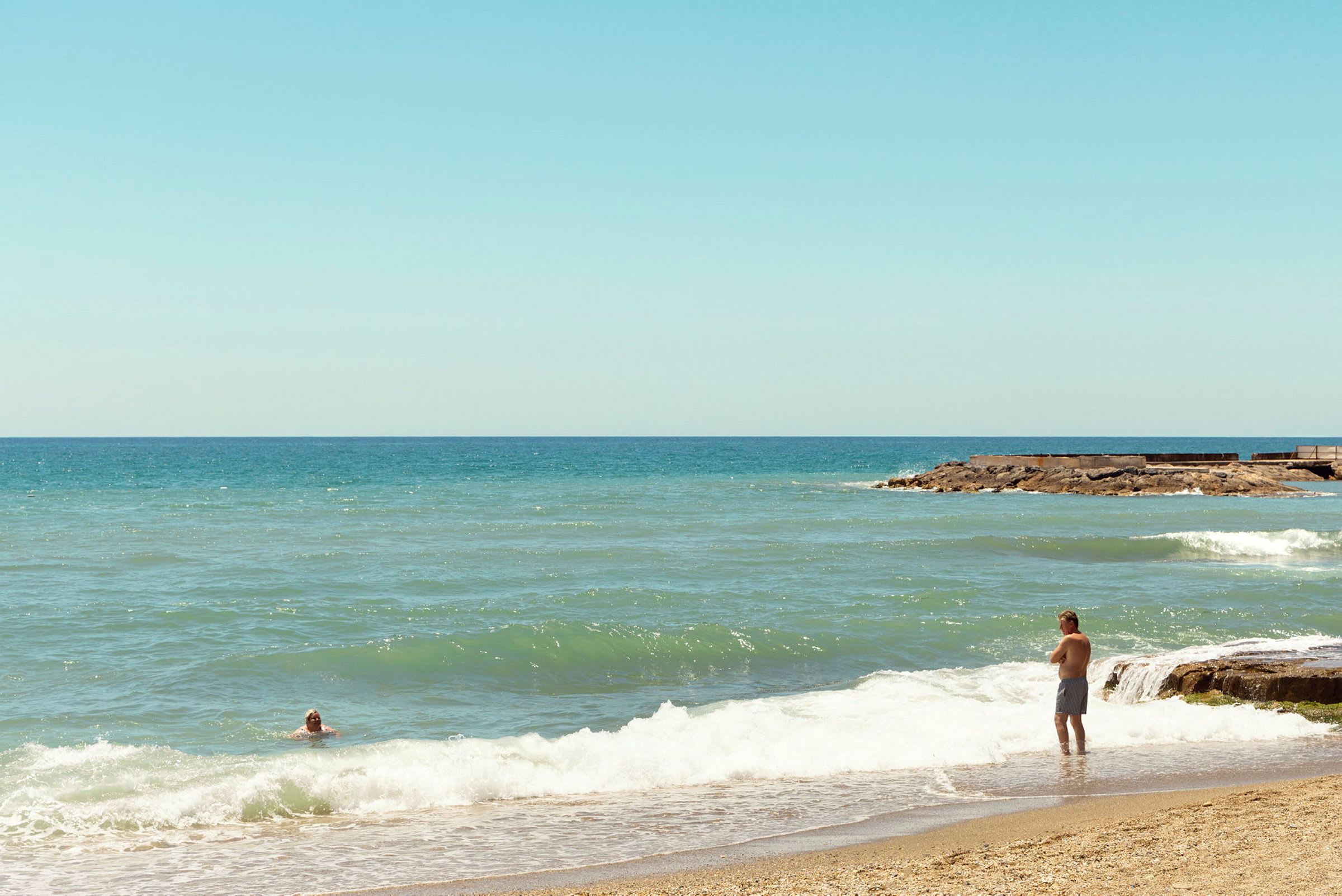 En strand med sand i förgrunden och havsvågor som rullar in. Två personer syns i bilden, en står vid strandkanten och en simmar i vattnet. (AI-genererad text)