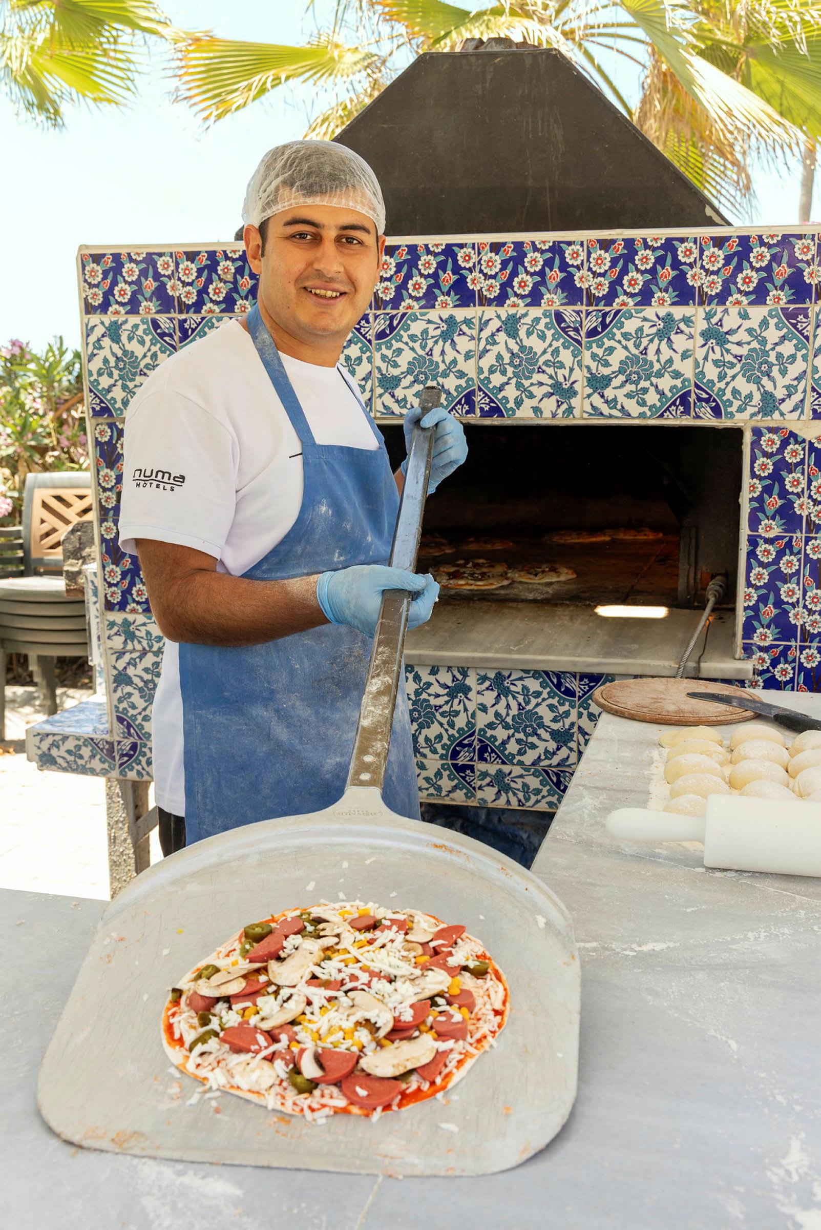 En pizzabagare håller en spade med en färdigförberedd pizza framför en ugn dekorerad med blå och vita kakelplattor. (AI-genererad text)