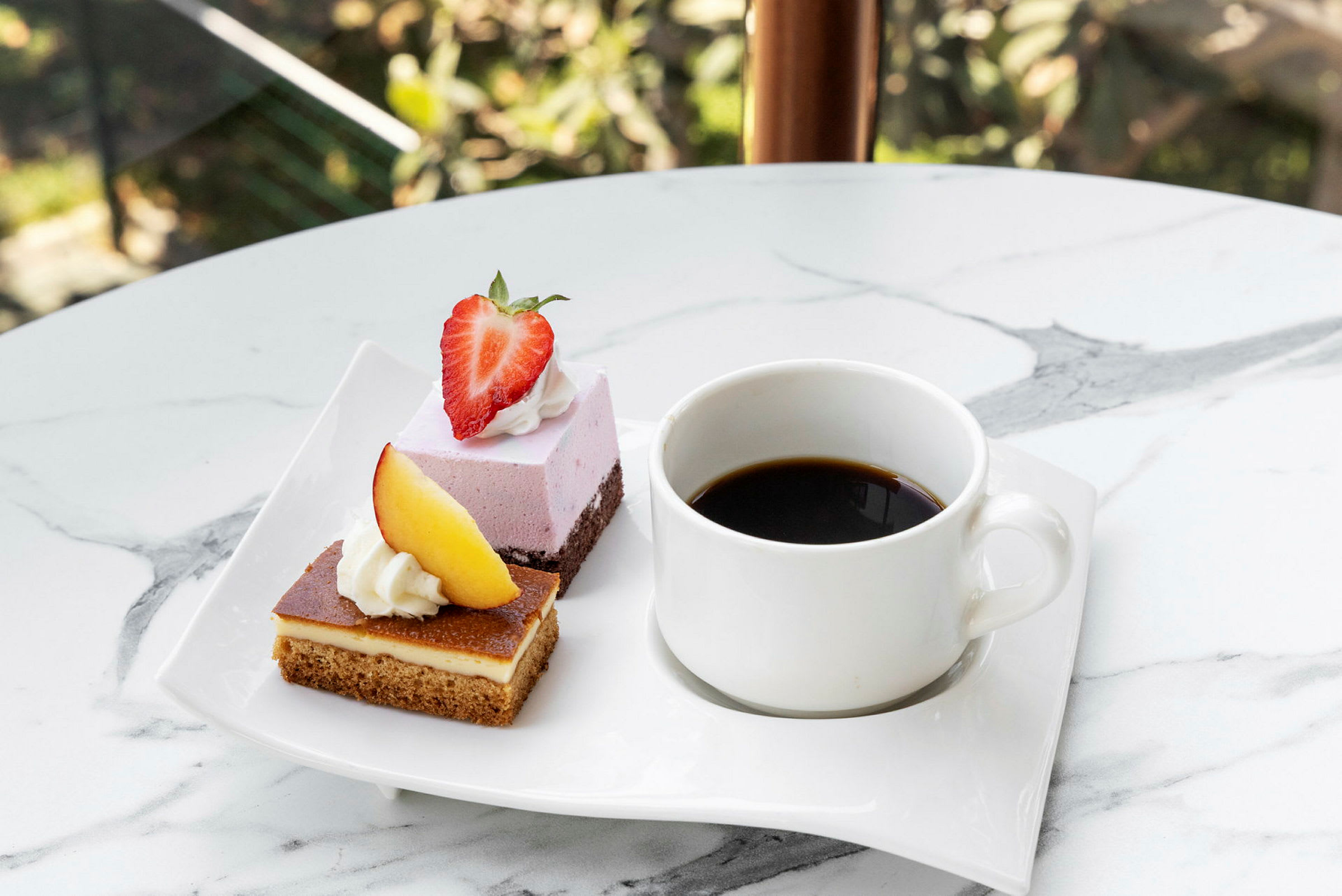 En vit tallrik med två desserter, en toppad med en jordgubbe och en med en bit persika, samt en kopp svart kaffe. (AI-genererad text)