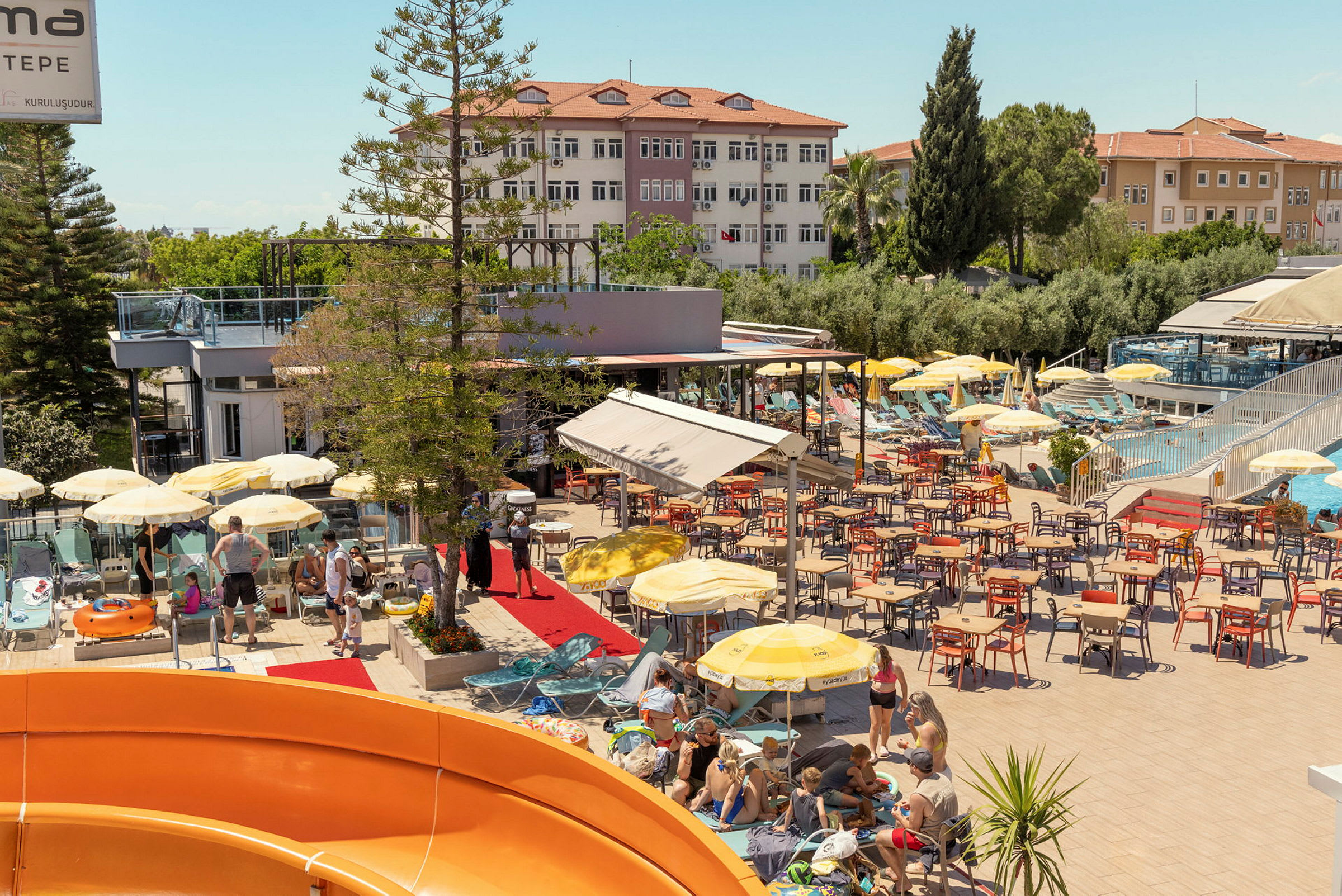 En utomhusområde med solstolar, parasoller och matbord, omgiven av byggnader och skuggande träd. (AI-genererad text)
