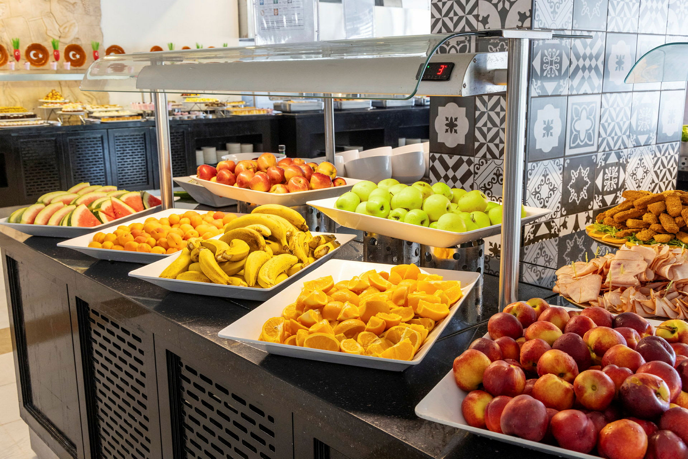 Ett buffébord med olika frukter som bananer, äpplen, apelsiner och vattenmelon samt kallskuret och andra tillbehör. (AI-genererad text)