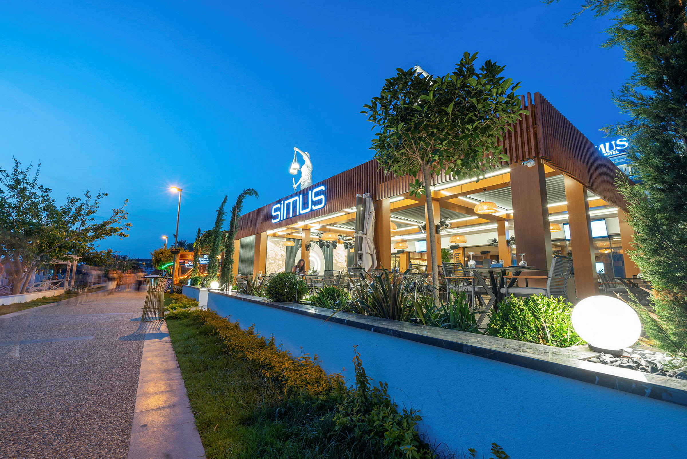 Modern restaurang med neonskylt, utomhusområde och växtdekorationer på en kvällsbelyst gata. (AI-genererad text)