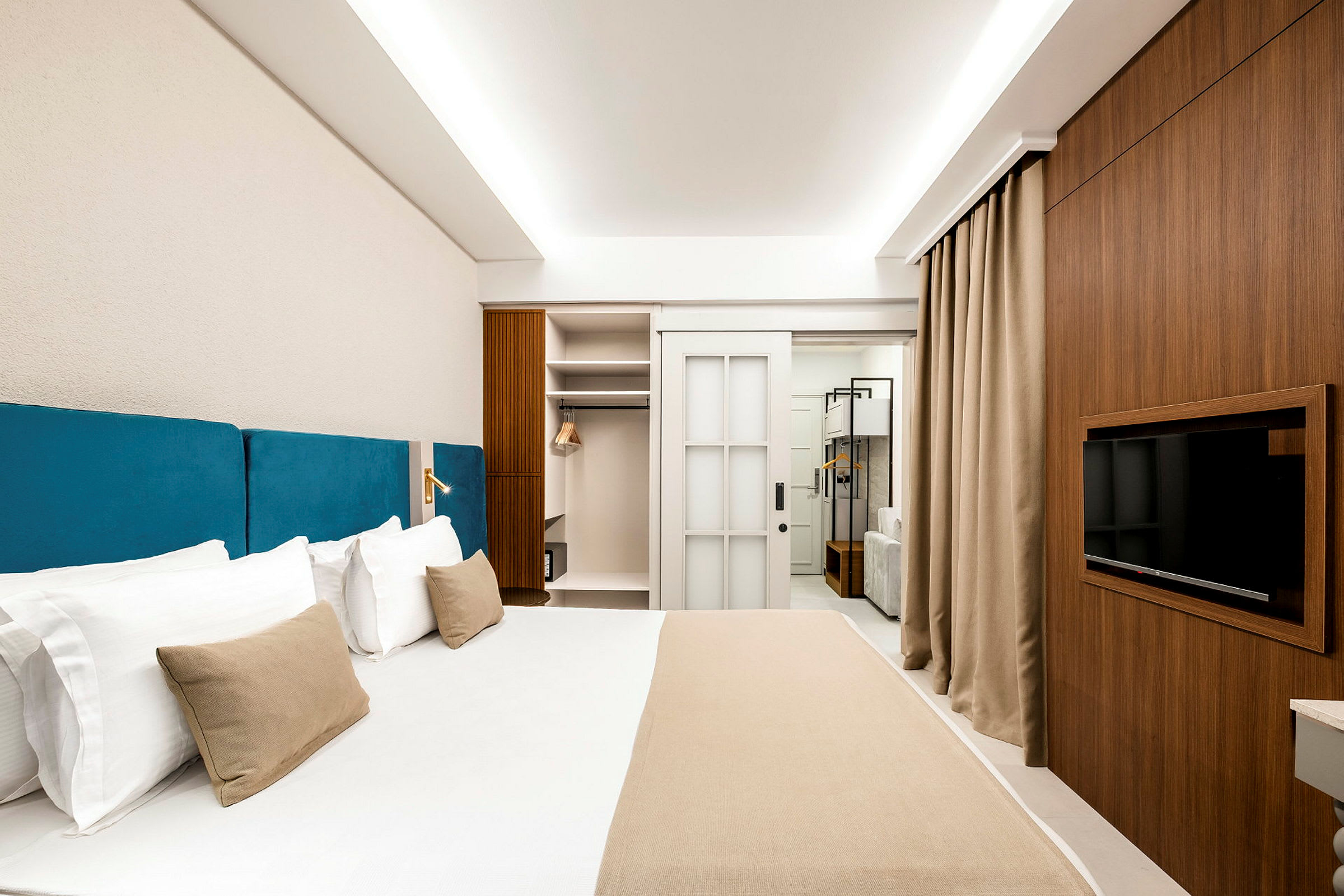 Hotellrum med en stor säng med blå sänggavel, en garderob, TV och stilren inredning. (AI-genererad text)