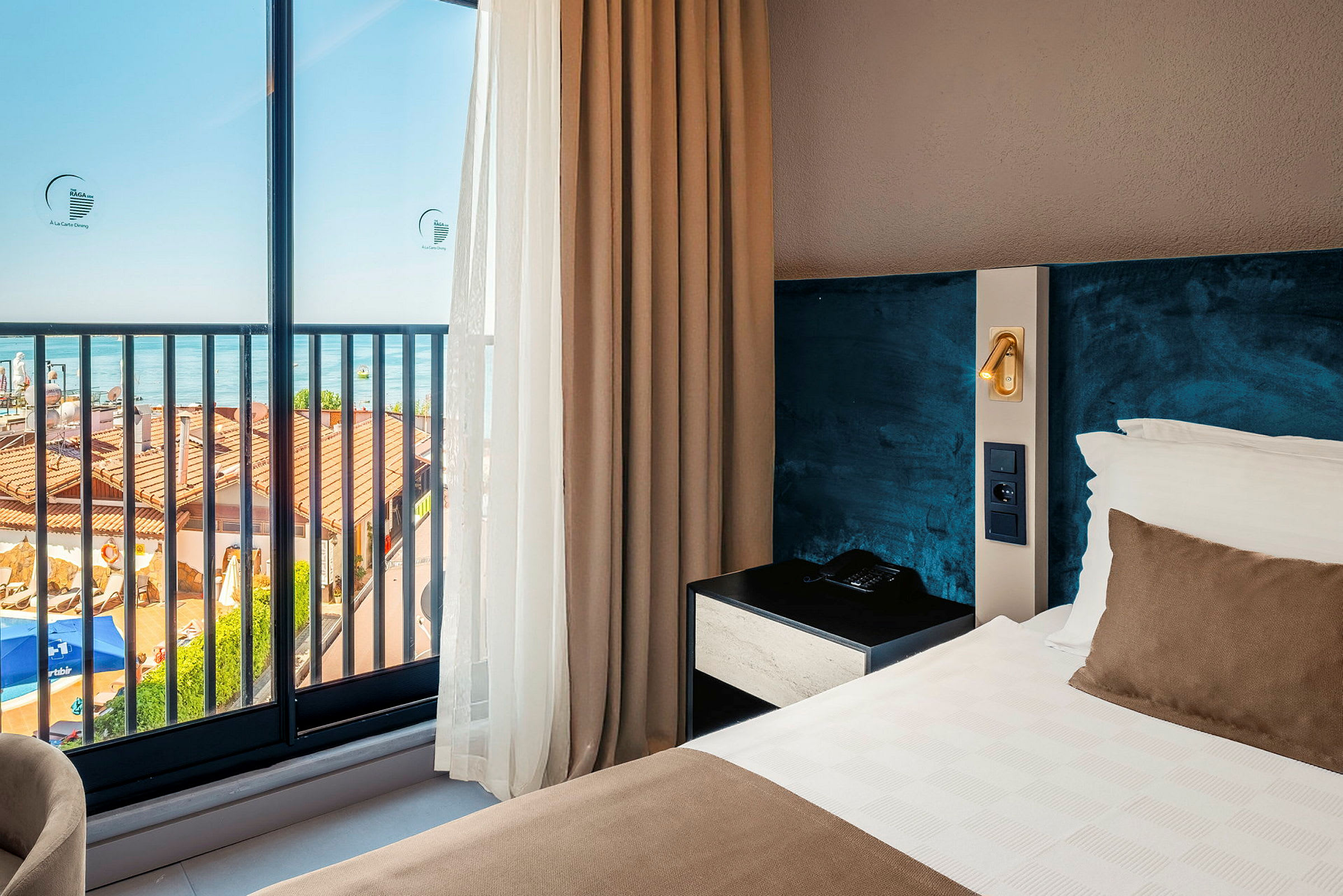 Ett hotellrum med havsutsikt genom stora glasfönster. Inredningen inkluderar en bäddad säng och ett sängbord. (AI-genererad text)