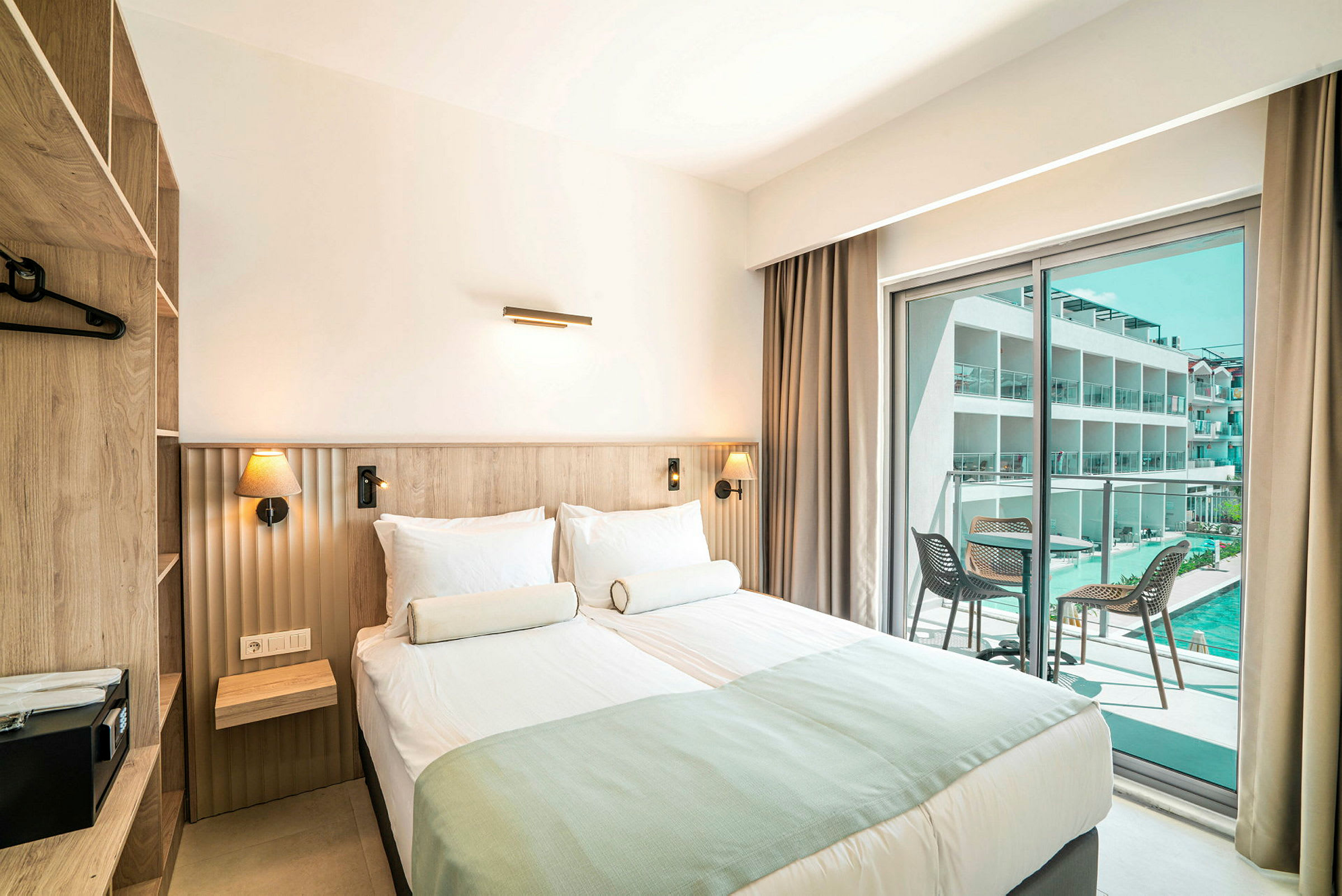 Ett hotellrum med dubbelsäng, modern inredning och glasdörrar som leder till en balkong med utsikt över en pool och byggnader. (AI-genererad text)