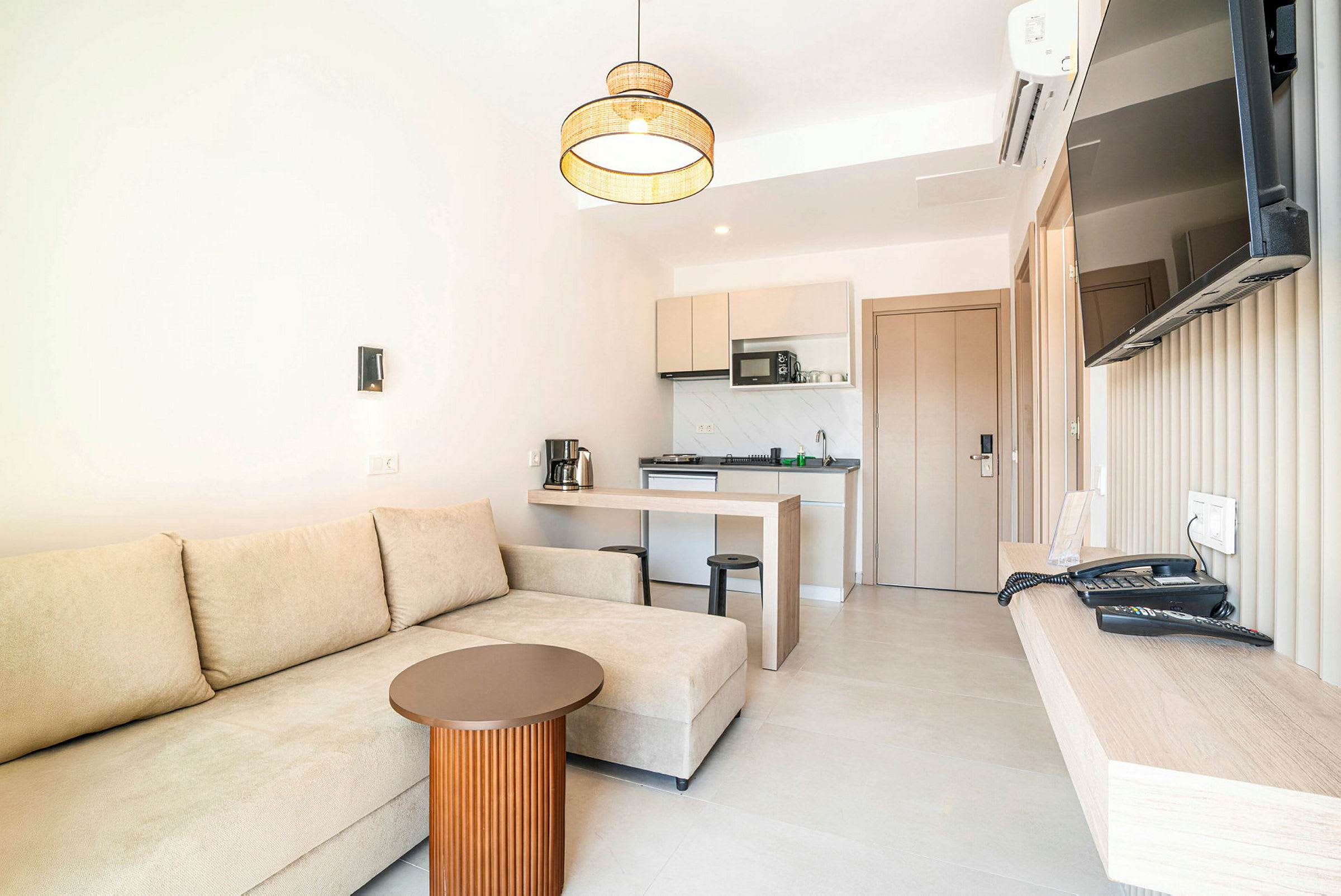 Ett hotellrum med en beige soffa, ett litet bord, ett kök och en väggmonterad TV. (AI-genererad text)