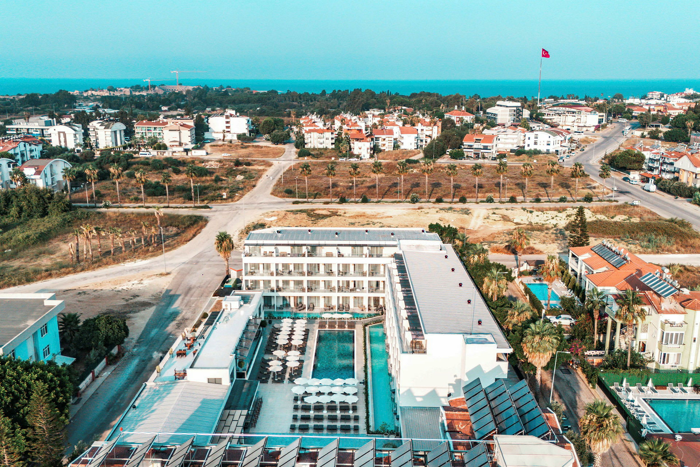 Flygfoto av ett hotell med poolområde, omgivande byggnader och en väg med palmer. (AI-genererad text)