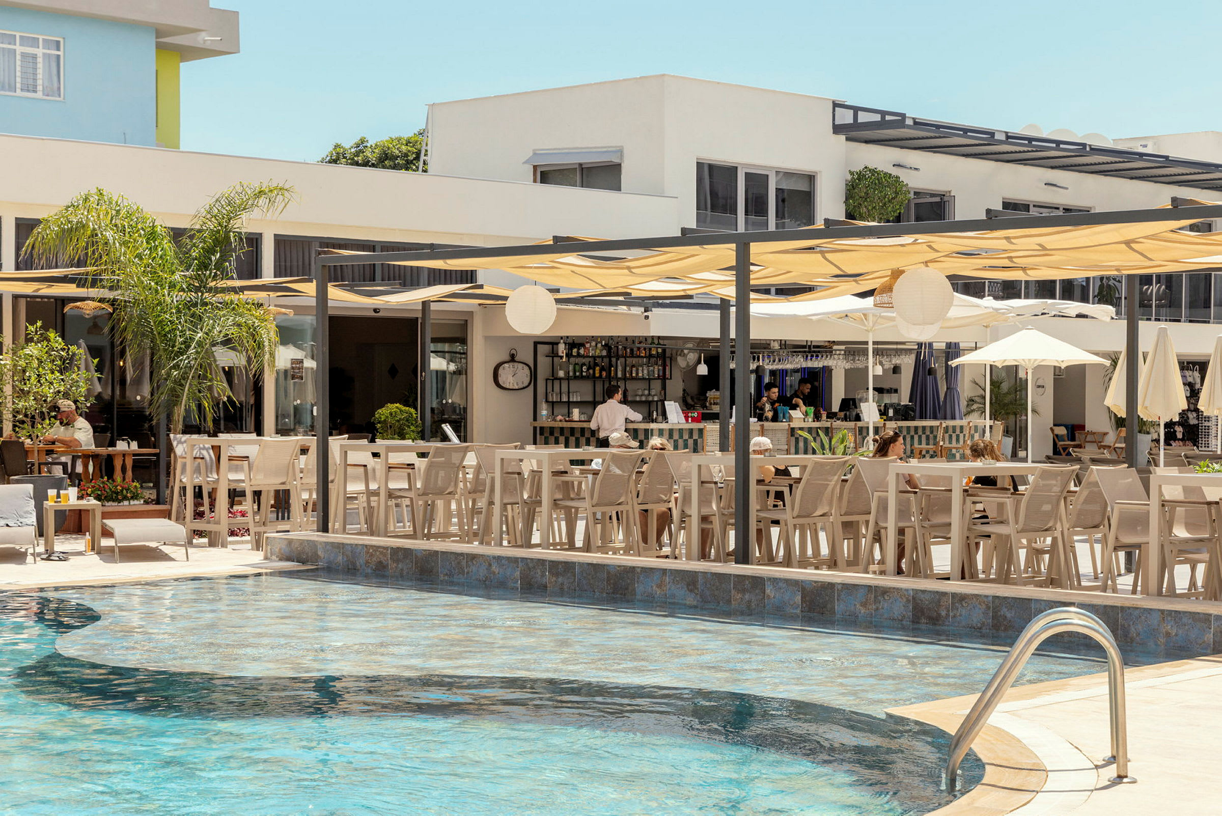 En utomhuspool med omgivande sittmöbler och parasoller vid en bar i bakgrunden. (AI-genererad text)