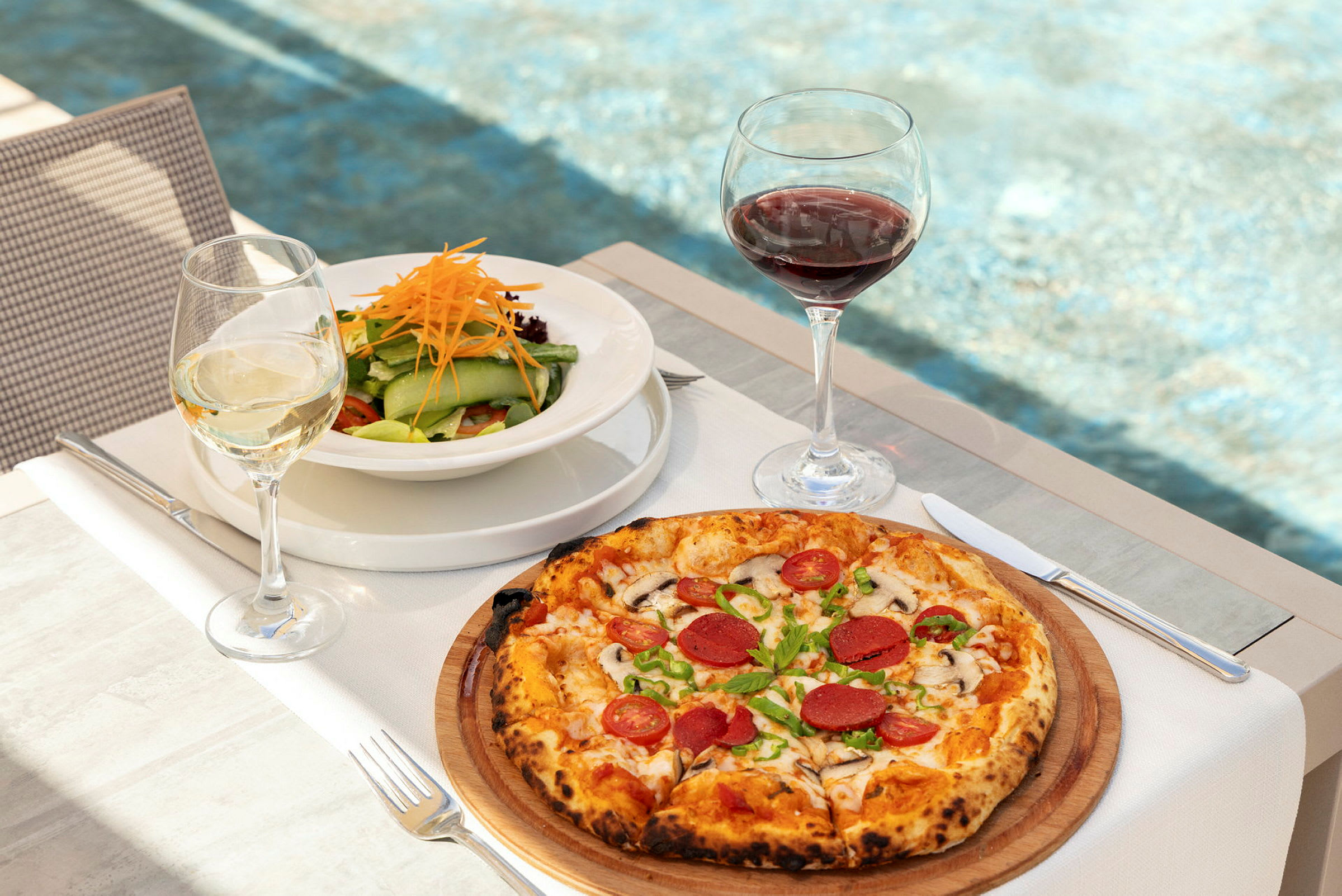 En tallrik med sallad och ett glas vitt vin bredvid en pizza med pålägg och ett glas rött vin, på ett bord med bakgrund av en pool. (AI-genererad text)