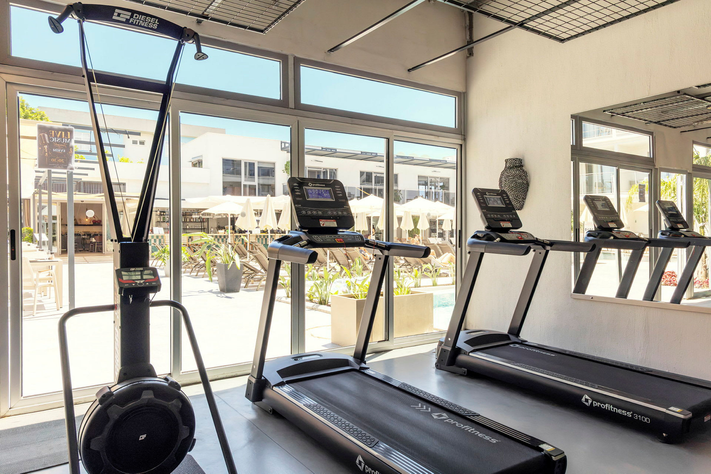 Ett gym med träningsmaskiner, inklusive löpband och en stakmaskin. Stora fönster visar utsikt över en terrass med parasoller. (AI-genererad text)