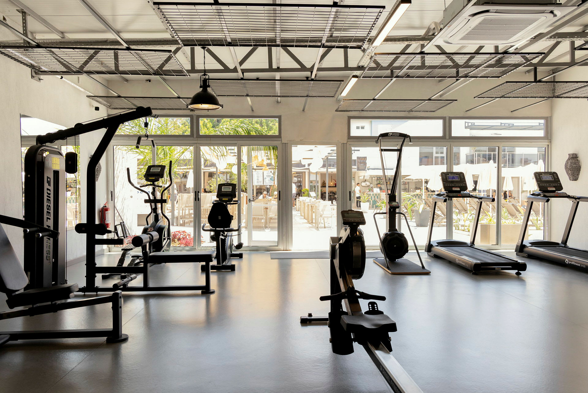 Ett gym med träningsutrustning inklusive löpband, motionscykel och roddmaskin, med glasväggar som visar utsikt över en uteplats. (AI-genererad text)