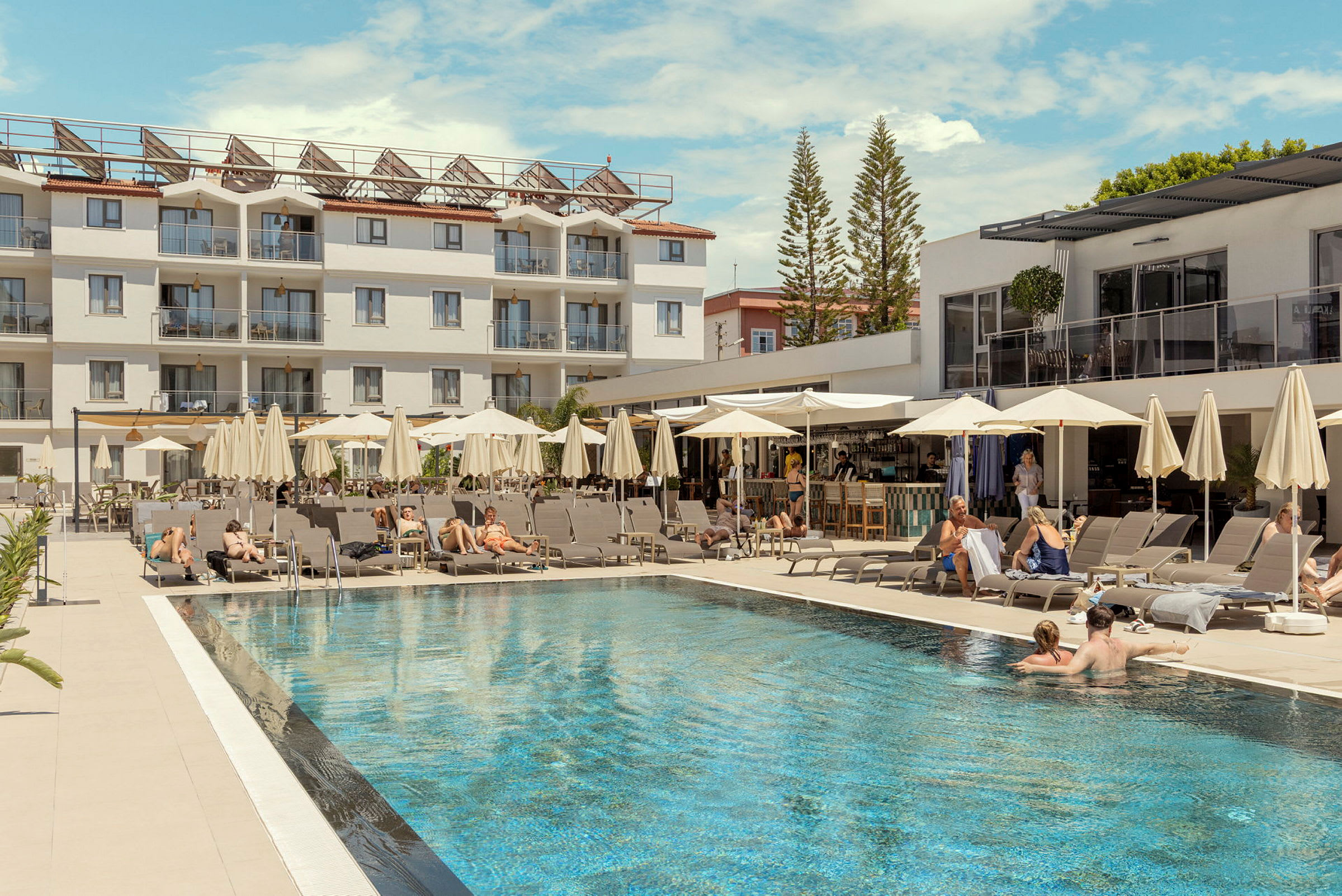 En hotellpool med solstolar och parasoll, omgiven av en vit byggnad och barer. (AI-genererad text)