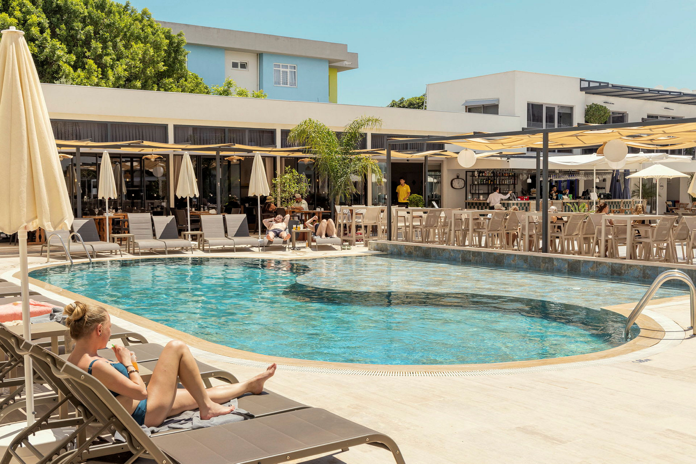 En utomhuspool omgiven av solstolar och parasoller, med en angränsande bar och sittplatser. (AI-genererad text)