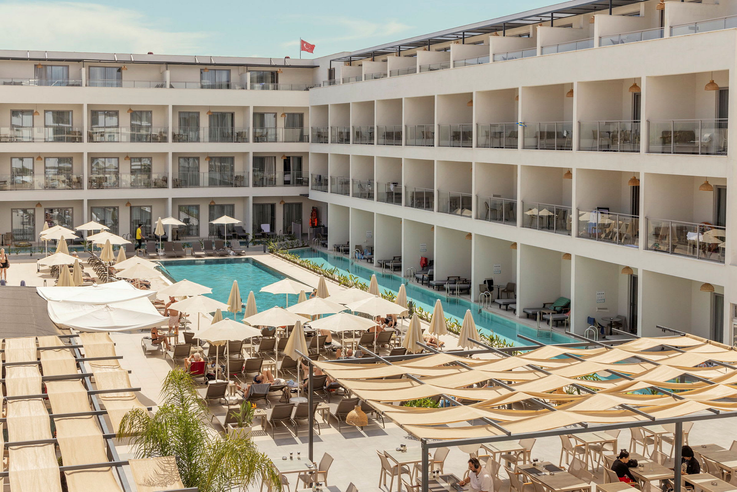 En hotellbyggnad med en pool i mitten samt solstolar och parasoller runtomkring. (AI-genererad text)