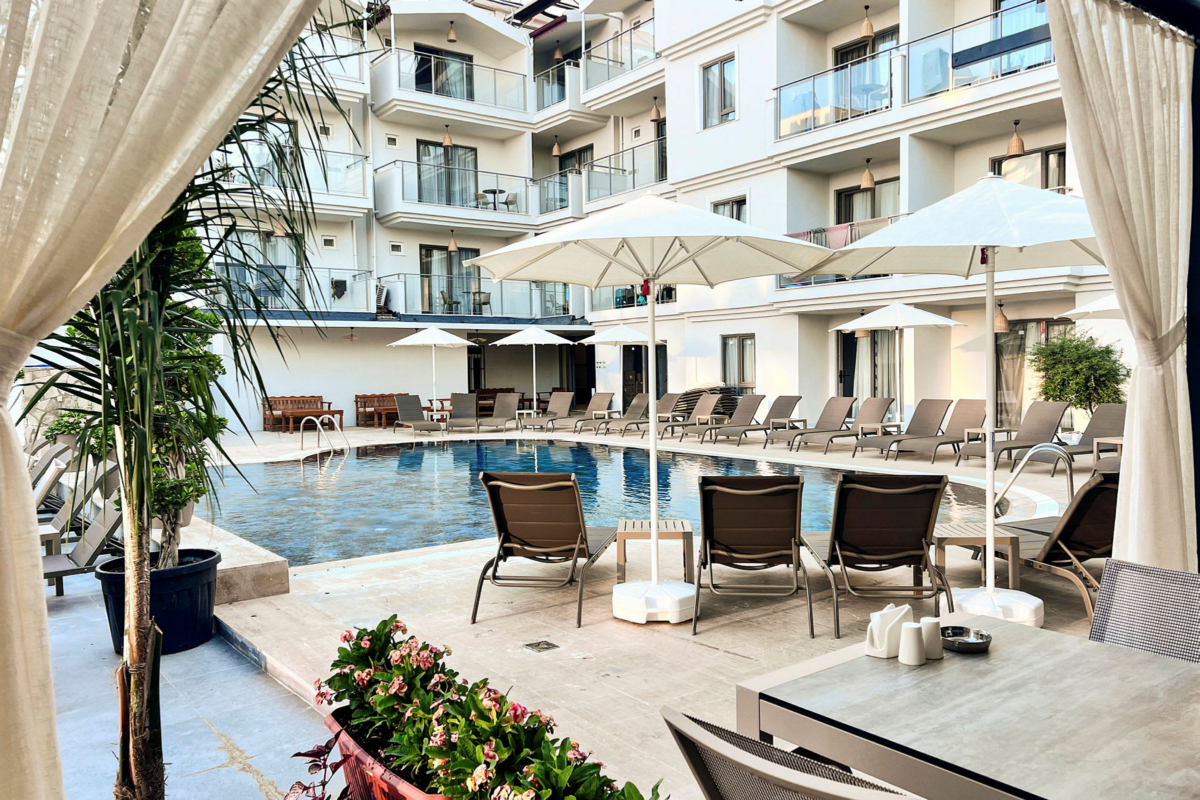 Bilden visar en hotellpool omgiven av solstolar, parasoller samt balkonger i bakgrunden. (AI-genererad text)