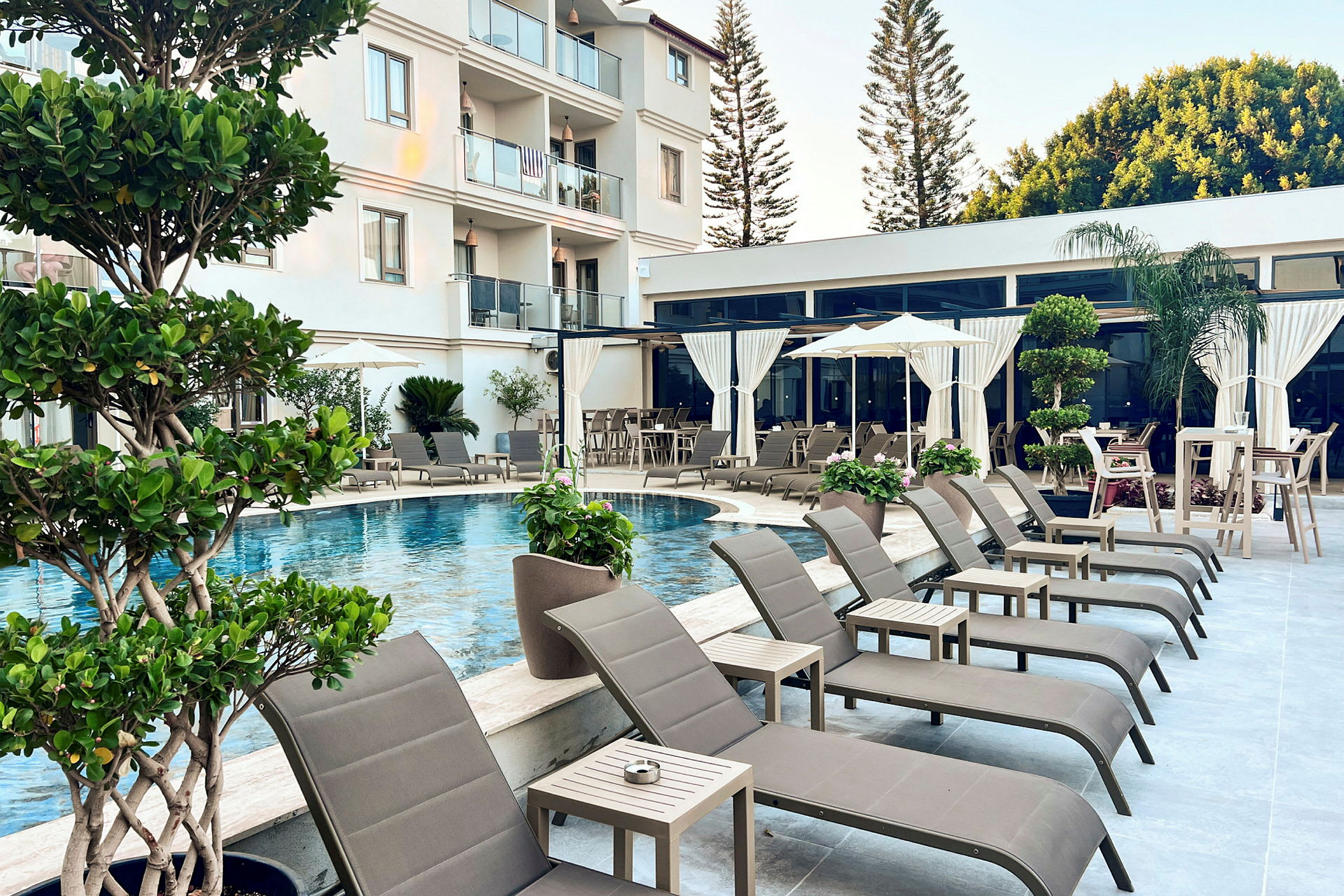 Poolområde med solstolar, parasoller, och bord samt omgivande växter vid ett hotell. (AI-genererad text)