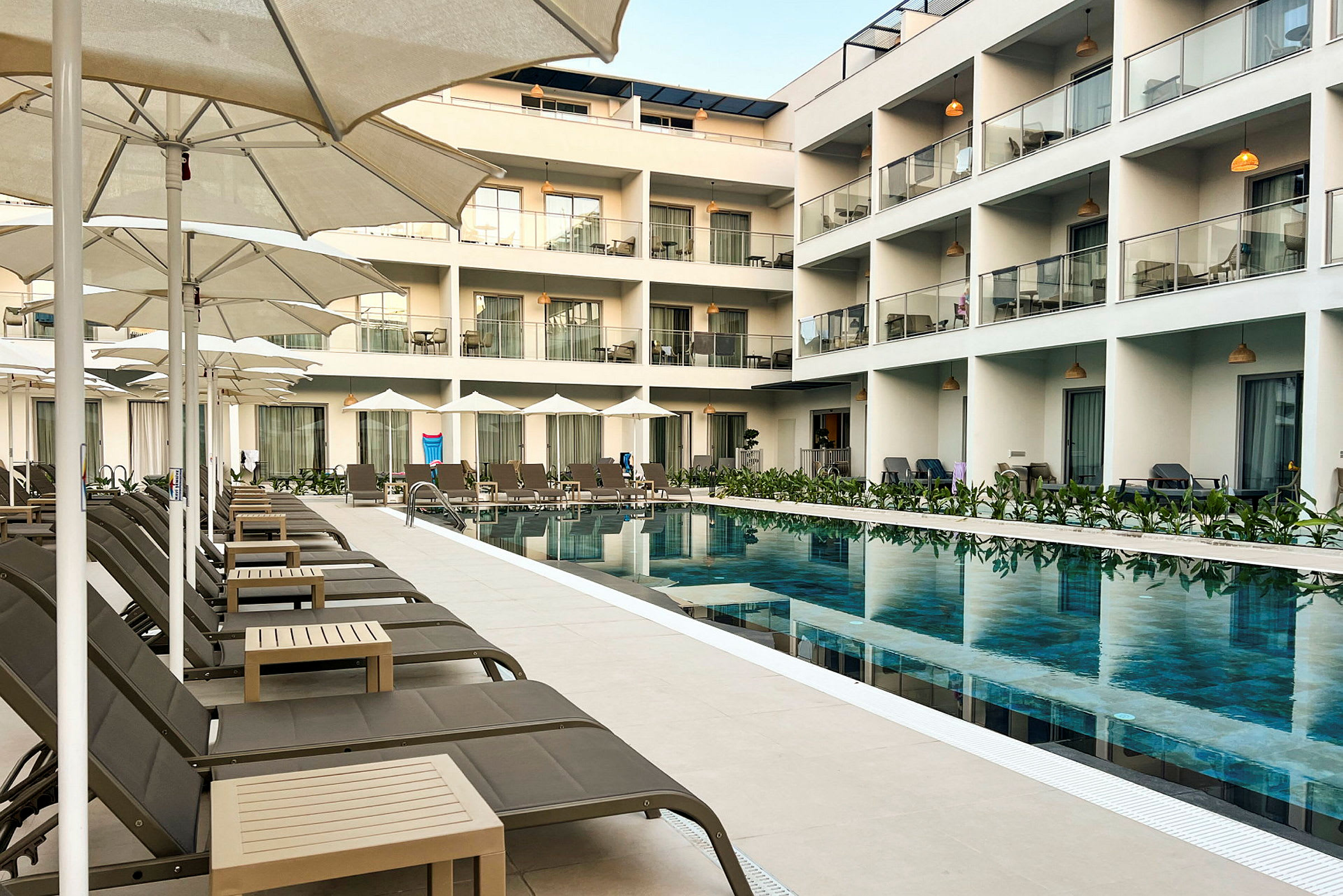 Ett hotell med en pool omgiven av solstolar och parasoller, samt balkonger ovanför. (AI-genererad text)